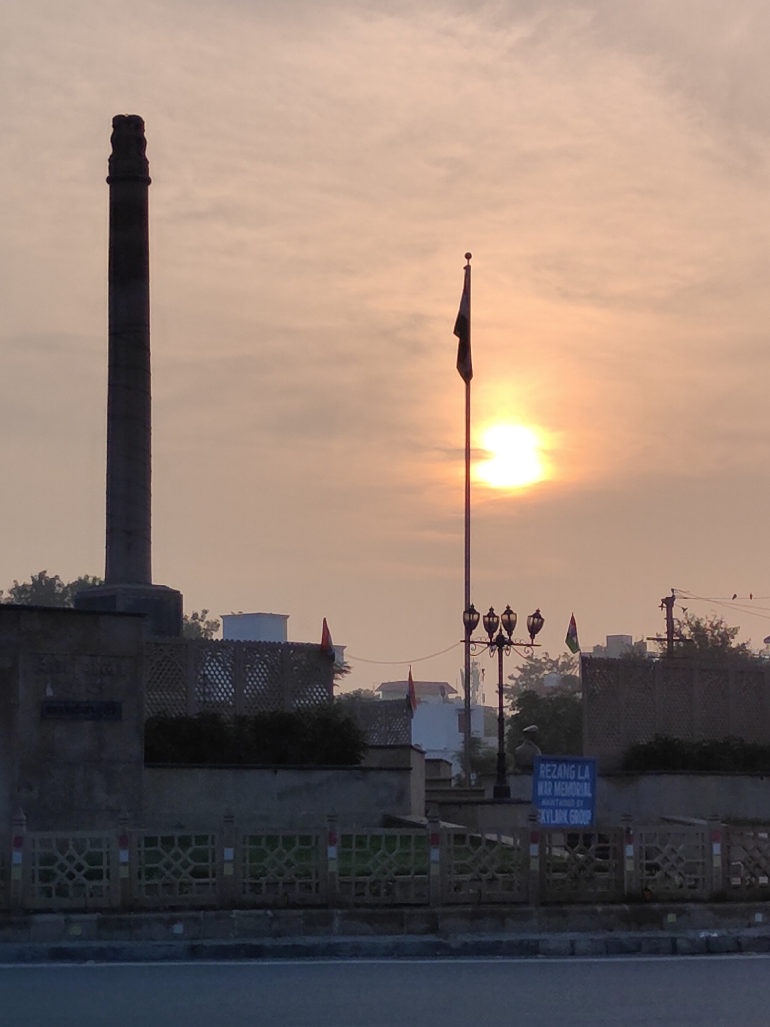 The image depicts a sunrise at the Rezag La War Memorial in Gurgaon, India. The memorial features a tall, dark pillar with an emblem at the top, likely the Ashoka Pillar, on the left side of the frame. A flagpole with a dark flag is positioned to the right of the pillar, with the sun prominently shining behind it. The sun appears as a bright, hazy orb, casting a warm glow over the sky, which is filled with pale clouds.
In the foreground, a road is visible, separating the viewer from the memorial complex. The memorial complex itself is enclosed by a decorative fence with a latticework design. Behind the fence, there are various structures, including buildings and trees. A blue signpost with white lettering is placed near the flagpole, indicating "REZANG LA WAR MEMORIAL MAINTAINED BY SKYLINK GROUP". Multiple lampposts are also present within the memorial grounds. Small flags, one red and one with green and white, are visible mounted on poles within the complex. The overall scene is serene, with the early morning light creating a peaceful atmosphere.