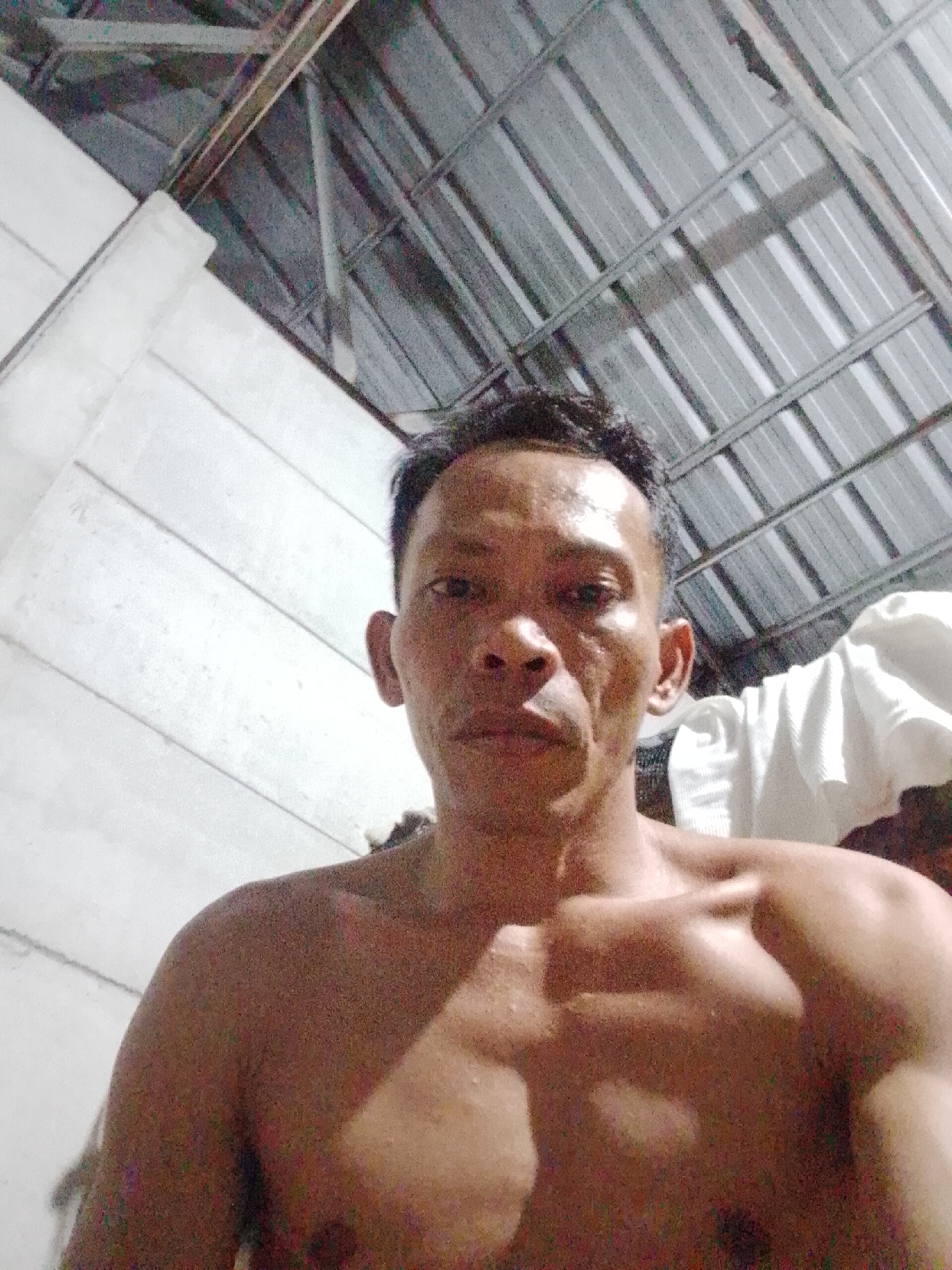 A shirtless man is centered in the frame, looking directly at the camera. The background features a white wall on the left and a metal corrugated roof overhead. The location is possibly Bukit Ketok, Indonesia.
