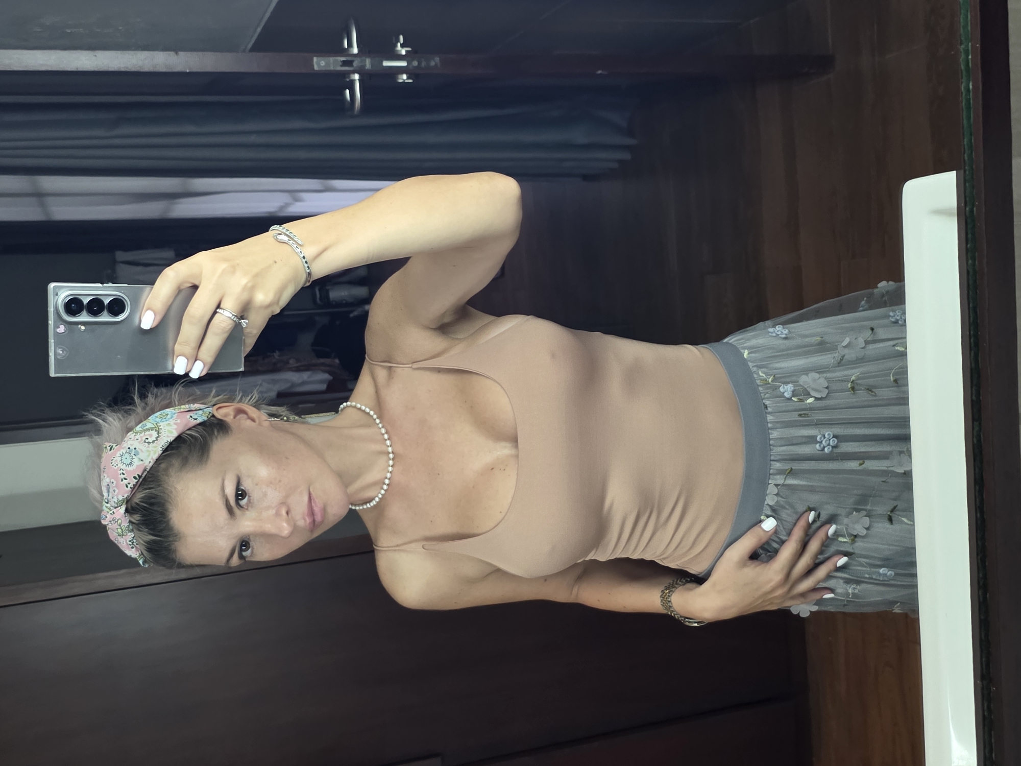 A woman is taking a mirror selfie in a room with dark wood paneling and a dark curtain. She is wearing a tan spaghetti strap top, a grey tulle skirt with floral embroidery, a pearl necklace, a beaded bracelet on her left wrist, and a patterned headband. She has white nail polish on her fingernails. Her hair is blonde and pulled back. She is wearing a ring on her left index finger. She is holding a silver smartphone in her right hand. The room appears to be a bedroom or dressing area. The overall impression is of a casual, personal moment.