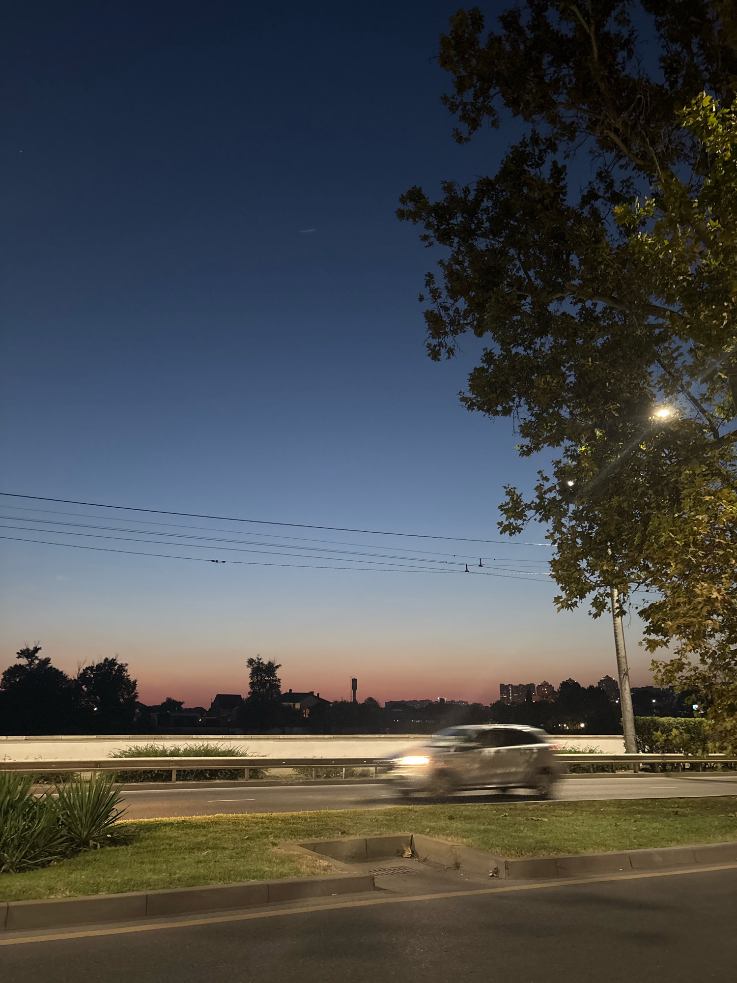 The image depicts a street scene at dusk in Krasnodar, Russia. The sky transitions from a deep blue at the top to a soft orange and pink hue near the horizon, indicating the end of daylight. Power lines stretch across the middle ground, supported by poles. On the right, a large tree with green and yellowing leaves is illuminated by a streetlamp, casting light rays through its branches.
In the mid-ground, a highway is visible with a central barrier and a moving vehicle, blurred due to motion, indicating traffic. Beyond the highway, a line of silhouetted trees and buildings marks the city's edge, with some taller structures visible in the distance. The foreground shows a grass verge with a curb and asphalt road. The overall atmosphere is calm and transitional, capturing the shift from day to night in an urban environment. The specific location within Krasnodar is not identifiable from the visual information provided.