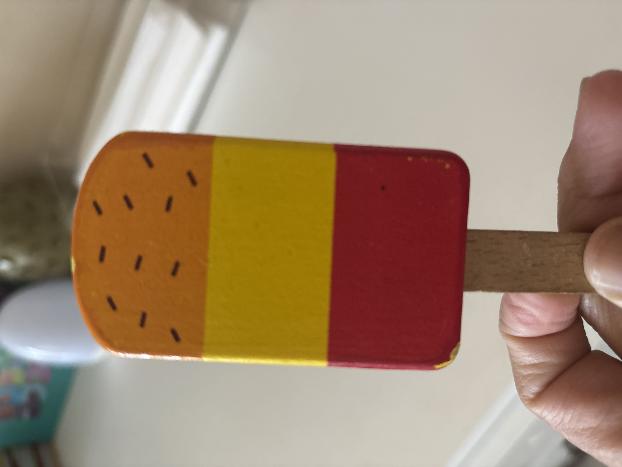 A hand is holding a wooden popsicle. The popsicle has a rounded end and is divided into three colored sections: orange with small black dashes, yellow, and red. The background is out of focus and appears to be indoors with muted lighting. The hand holding the popsicle suggests the object is being presented or played with. The image does not contain any visible text.