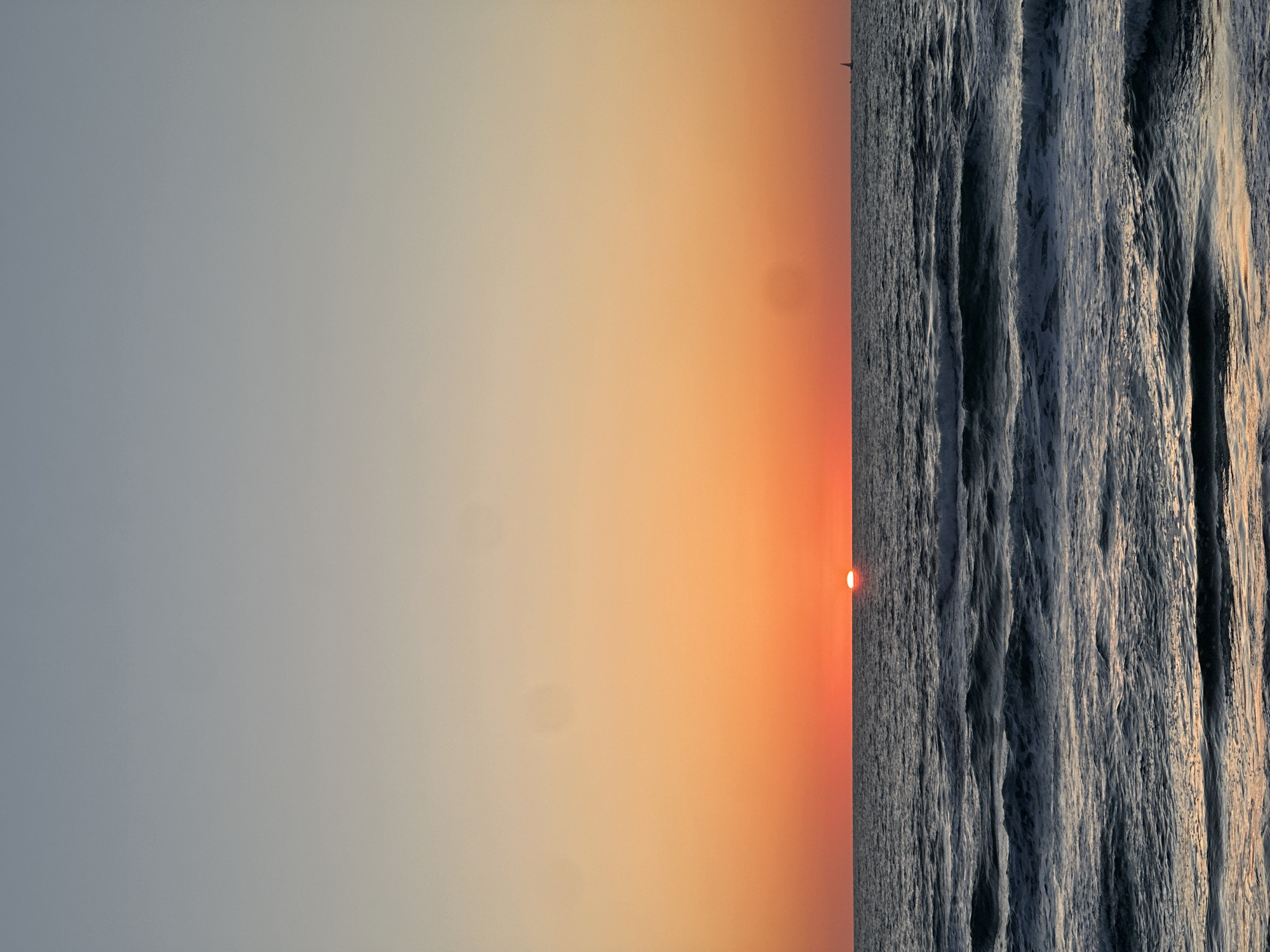 The image depicts a tranquil sunset over the sea, with the horizon line spanning horizontally across the mid-section of the frame when correctly oriented. The scene is likely along the coast of Tel-Aviv, Israel, overlooking the Mediterranean Sea.
The sky, occupying the upper portion of the image, displays a beautiful gradient of warm hues. It transitions from a brilliant orange and deep red directly above the horizon, where the sun is positioned, gradually fading into lighter shades of peach, pale yellow, and a soft, desaturated blue-grey towards the top. The setting sun is visible as a small, intense red-orange circle resting precisely on the horizon line.
Below the horizon, the sea is dark and expansive, characterized by numerous visible waves that create a textured surface. The undulating water reflects some of the last golden light from the sun, highlighting the crests of the waves with subtle glimmers. A faint, dark silhouette, possibly a distant boat or a navigational marker, can be seen on the horizon line against the glowing sky.
The overall atmosphere is one of serene natural beauty as day transitions to night. There is no visible text in the image.