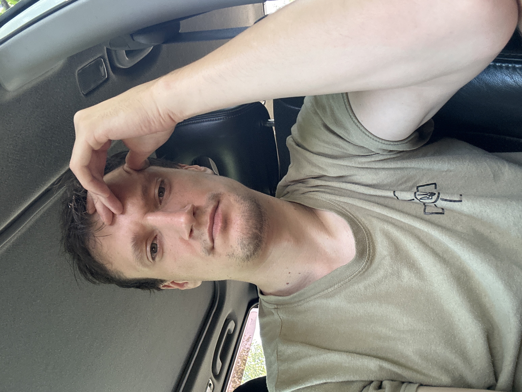 A man with short, dark, messy hair and a light beard is sitting in the driver's seat of a vehicle. He is wearing a tan t-shirt with a black emblem on the left side of his chest. His right hand is raised to his forehead. The man's eyes are open and he is looking towards the camera. The interior of the vehicle is visible, including the dashboard, sunroof, and the back of a black headrest. Outside the vehicle, foliage can be seen through the windows. The image appears to have been taken during the daytime, with natural light illuminating the scene. The man's posture and facial expression suggest he may be experiencing discomfort or deep thought.