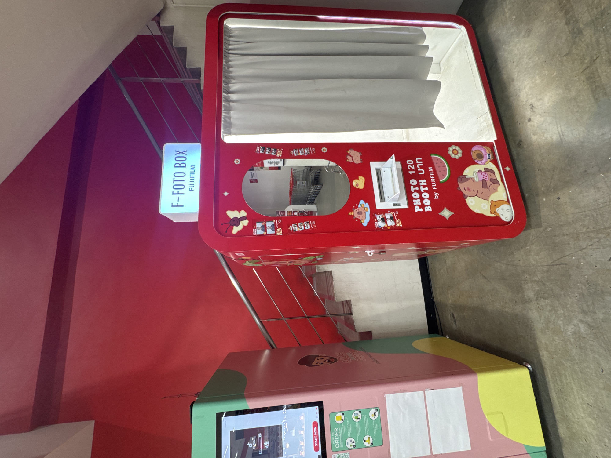 This image captures an indoor scene featuring two colorful photo booths, likely in a commercial or public area in Bangkok, Thailand, indicated by the Thai word "บาท" (Baht) visible on one of the machines.
The primary subject is a vibrant red photo booth positioned prominently on the left side of the frame. This booth, branded "F-FOTO BOX" and "FUJIFILM," features a curtained section at the top where users would sit for photos. Below this section is a reflective surface or screen, with the text "PHOTO 120 BOOTH บาท by FUJIFILM" prominently displayed. The red exterior is adorned with various cartoon-like stickers, including a bear, watermelon, donut, and bee, creating a playful aesthetic. A glowing sign above the booth reiterates "F-FOTO BOX FUJIFILM."
To the right and slightly below the red booth is a second, larger photo booth with a distinctive pastel color scheme of pink, green, and yellow. This booth features a large digital screen displaying a user interface with a "START NOW" button and the "Tik Tok" logo, suggesting advanced photo or video capabilities. Instructions titled "HOW TO ORDER" are also visible on its side, along with the partially obscured word "ANGRY," possibly part of a game or branding.
The background includes a red wall on the left and a visible staircase with white steps and silver railings on the right, implying a multi-level structure. The floor is made of concrete. No people are visible in the image, but the presence of the booths clearly indicates a space designed for leisure and entertainment, where people can take instant photos or create digital content. The overall lighting appears to be artificial and bright, typical of an indoor commercial environment.