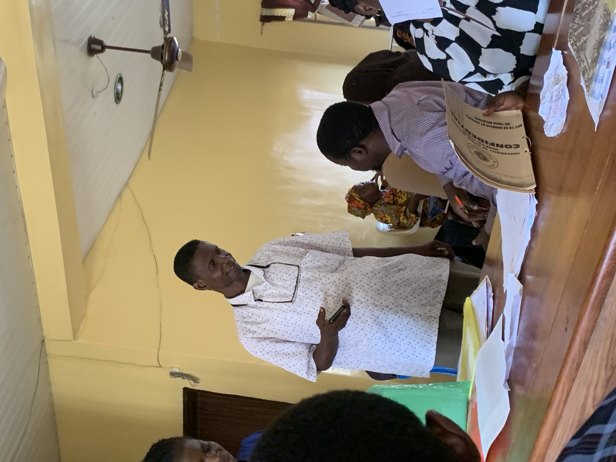 The scene depicts a meeting or presentation occurring indoors, likely in the city of Oko Erin, Nigeria. A man wearing a white patterned short-sleeved shirt stands addressing a group of people seated at a wooden table. He holds a mobile phone in his left hand and gestures with his right. Another person is at the table and is looking at a man with documents and a confidential folder. Other individuals are partially visible around the table. A ceiling fan hangs from the ceiling above. Documents and papers are visible on the table. The walls are painted yellow.