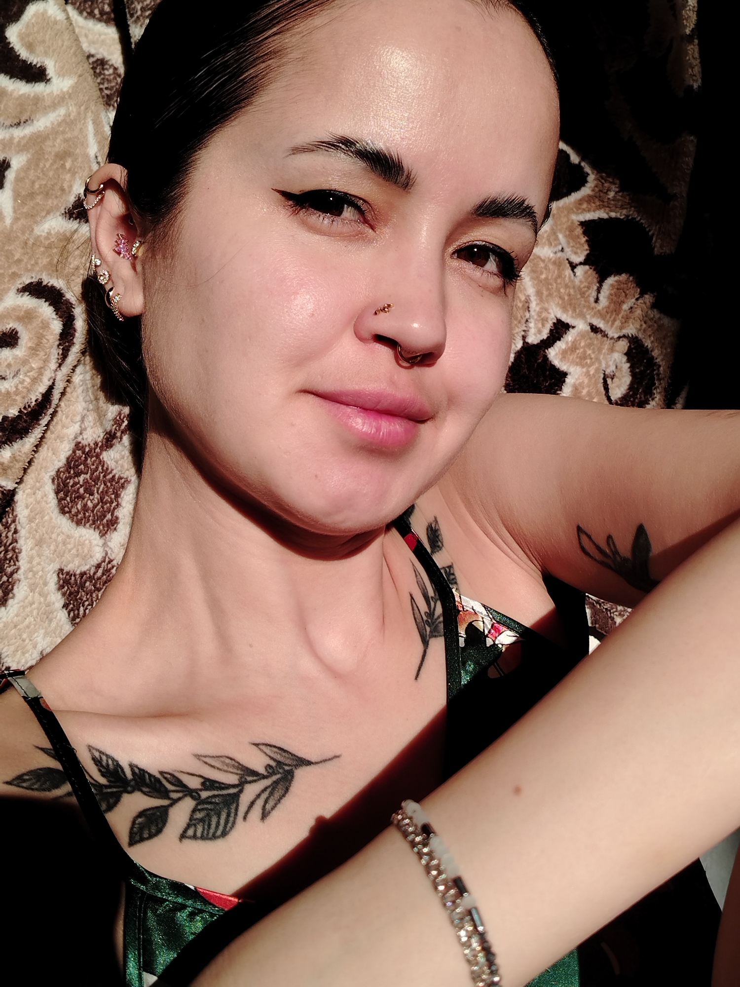 A close-up, high-angle selfie shows a fair-skinned person with dark hair pulled back, revealing multiple piercings in their left ear and a nose ring. The person is wearing what appears to be a green and red floral-patterned camisole. Visible tattoos of branches or leaves are on the chest and upper arm. A bracelet adorns the wrist of the visible arm. The background is a patterned brown and beige fabric. The person is facing the camera. Location details: Unknown, Unknown.