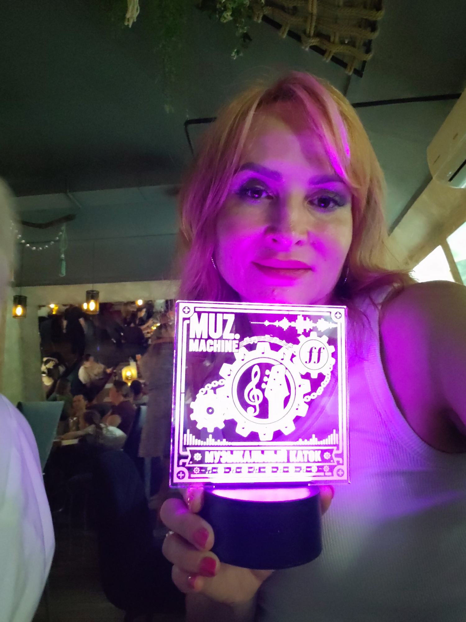 A woman with blonde hair and pink highlights holds a lit square acrylic object with a black base in front of her. The object displays a white graphic with the text "MUZ. MACHINE" at the top, a treble clef inside a gear and a guitar headstock graphic in the center, and "МУЗЫКАЛЬНЫ KATOK" at the bottom, along with other abstract designs. The object is illuminated with purple light, casting a purple glow on her face and chest. She is wearing a light-colored tank top. In the background, a dimly lit room is visible with several people seated at tables, suggesting a restaurant or bar environment. The woman's hands are visible, holding the object with her fingers extended. The overall lighting suggests an indoor, possibly evening setting.