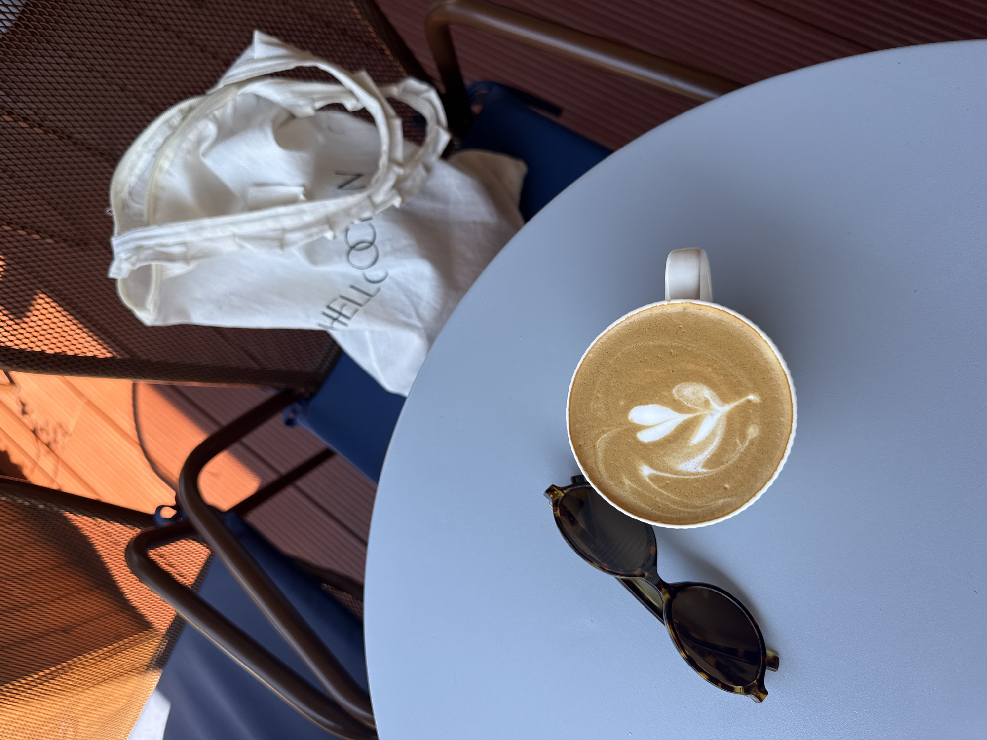 A top-down, medium shot shows a light blue, round table outdoors with a white cup of coffee with latte art, a pair of tortoise-shell sunglasses, and a white canvas tote bag with the text "HELLOCAN" on it. The table is set on a wooden deck, with the backs of two chairs visible in the background. One chair is made of brown metal tubing and a mesh material, and the other is blue. The scene is lit by warm, orange sunlight. The overall atmosphere is relaxed and casual. This scene is likely in Fethiye, Turkey, given the presence of the "HELLOCAN" tote bag, a local brand.
