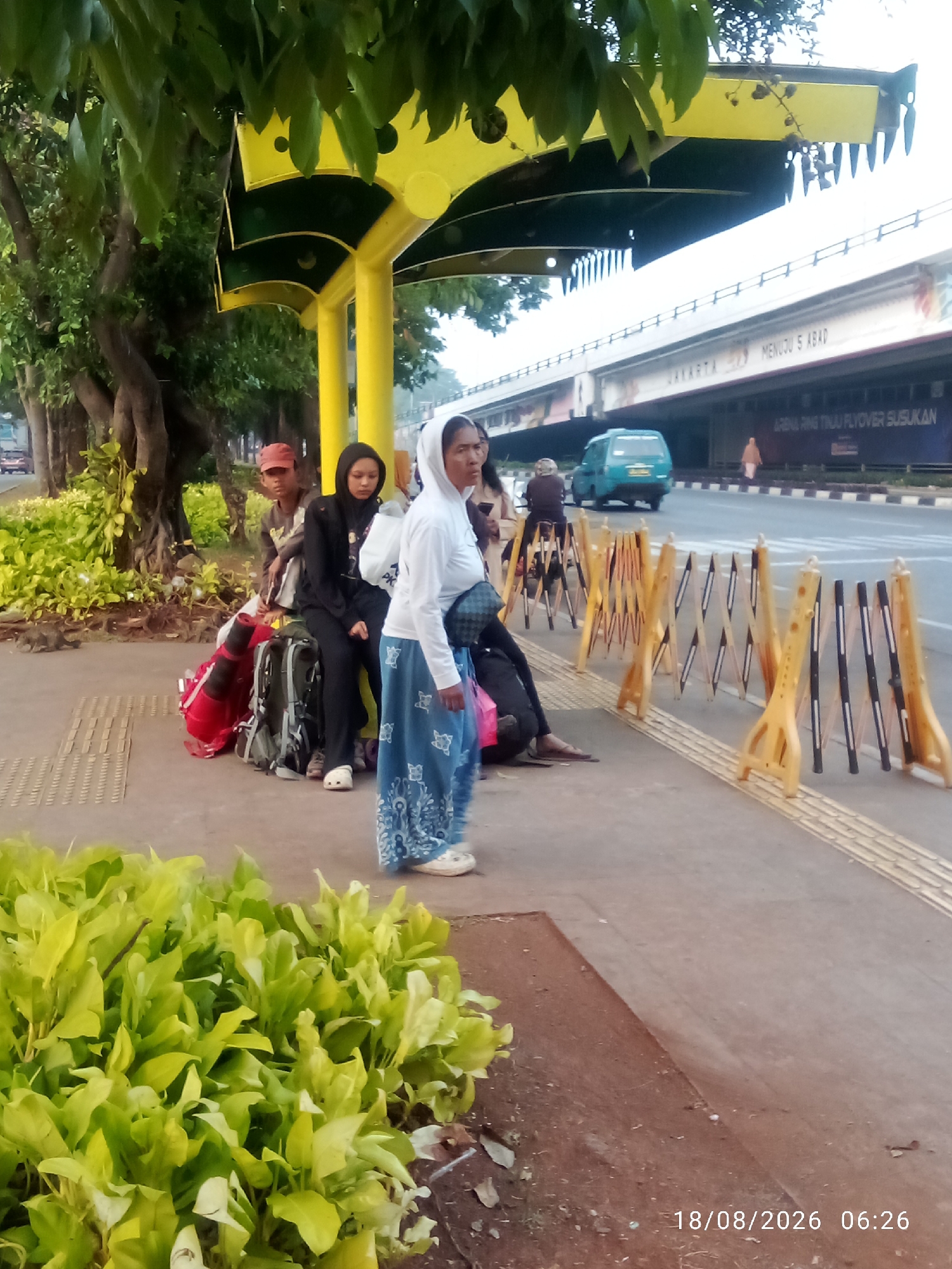 This image depicts a street scene in East Jakarta, Indonesia, likely near a bus stop or waiting area, as indicated by the presence of people sitting with luggage under a covered structure, possibly a bus shelter. The structure's yellow and dark green color scheme and sawtooth-like trim suggest an urban design element. In the foreground, lush green foliage is visible, contrasting with the paved walkway and reddish-brown soil.
Several individuals are present. On the left, a young person wearing a red cap is seated, while next to them, a young person in a black hoodie and hijab is also seated. They are accompanied by luggage, including a red duffel bag and several backpacks. To the right, a woman in a white hoodie and a blue patterned skirt stands, facing away from the camera.
In the background, a multi-lane road is visible, with a turquoise van in motion. An elevated highway structure with advertisements is also prominent. Barriers, likely for crowd control or traffic management, are positioned along the walkway. The overall lighting suggests it is daytime, possibly early morning given the timestamp at the bottom right of the image: 18/08/2026 06:26. The environment appears to be an outdoor public space within a city, with trees lining the walkway and a busy road indicating urban activity.