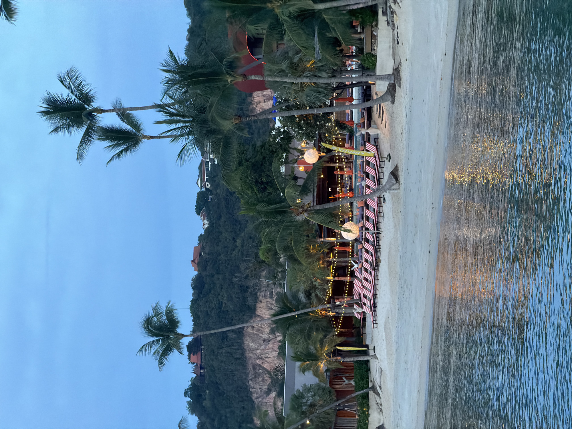 This image depicts a tranquil beach scene in Baan Bang Rak, Thailand, likely during twilight. The foreground is dominated by calm, blue water reflecting scattered golden lights from the shore. A sandy beach stretches beyond, lined with numerous red and white striped lounge chairs, suggesting a resort or public beach area. Palm trees are scattered throughout the scene, with some reaching towards the pale blue, cloudless sky. In the background, a lush green hillside is dotted with several red-roofed buildings, possibly villas or resort accommodations. String lights and large spherical lanterns illuminate the beach area, creating a warm and inviting atmosphere. The overall impression is one of peaceful relaxation and tropical beauty at the end of the day.