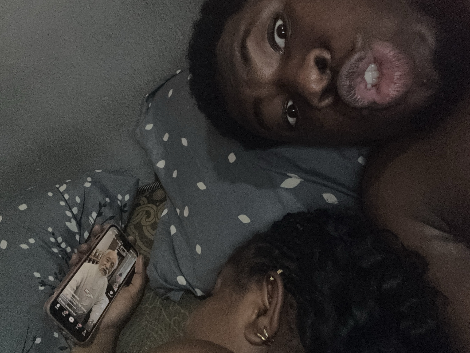 A man and woman lie in bed, the man looking into the camera with pursed lips. The woman’s head is on a pillow with white floral design. A hand holds a cell phone displaying a TikTok video of an older white man. The setting is a room with a gray wall. The location is Gbongan, Nigeria.