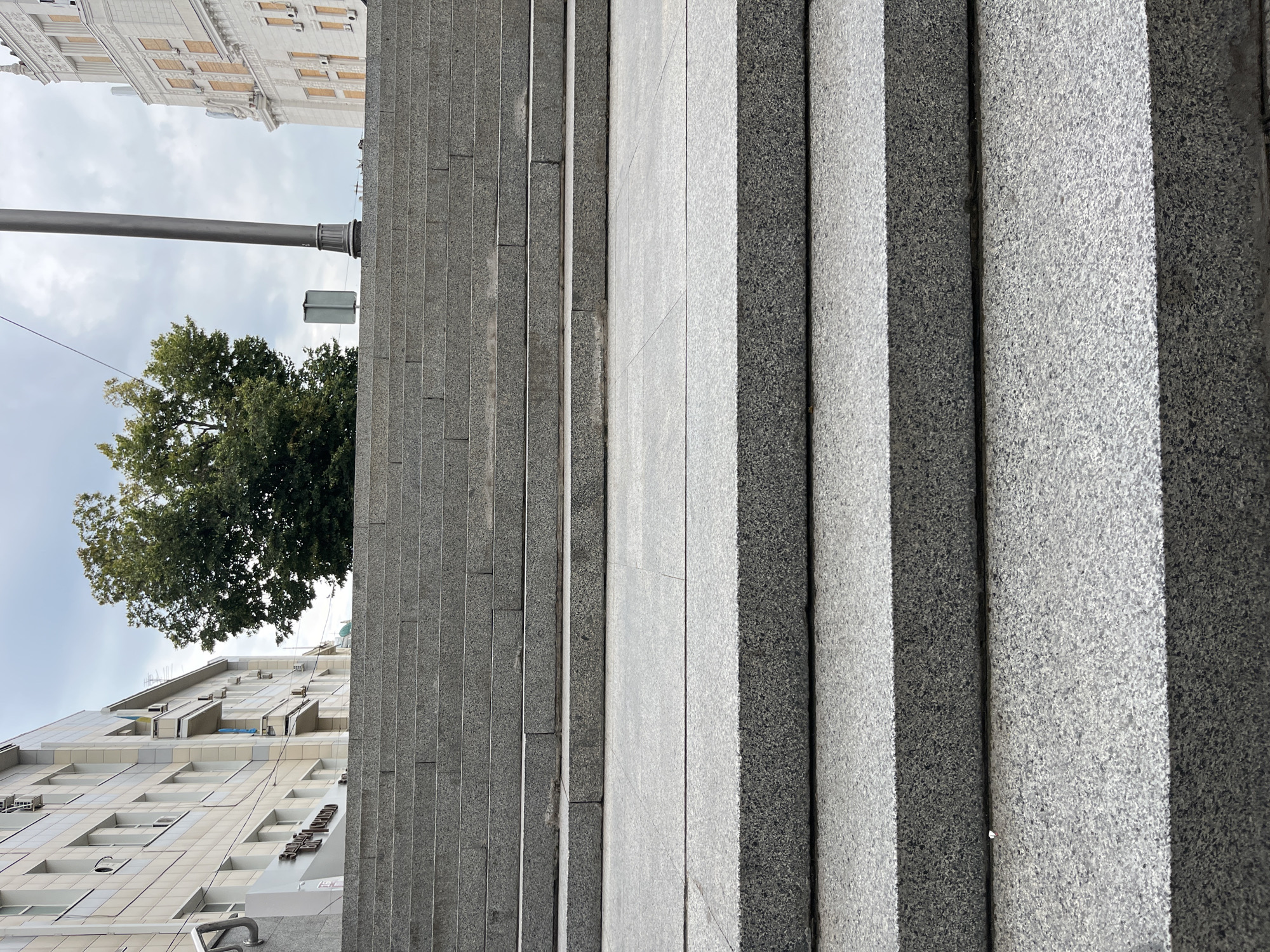 The image depicts a wide, outdoor granite staircase in a city. The stairs are constructed from large, polished granite blocks, with a speckled gray and black texture. The perspective is from the bottom of the staircase, looking upwards.
In the background, several buildings are visible. To the left, a modern, multi-story building with a light-colored facade and numerous windows is partially visible. A sign with Cyrillic lettering is mounted on the lower part of this building. To the right, a more ornate, taller building with cream-colored facade and decorative elements can be seen. The sky is overcast and gray, with some light peeking through.
Between the buildings and above the staircase, a large, mature deciduous tree with green foliage stands prominently. A tall, slender metal lamppost is positioned to the right of the tree. A small, rectangular green sign is mounted on a post to the left of the lamppost, also in the background. The scene suggests an urban public space, possibly a plaza or the entrance to a significant building. The overall impression is one of civic architecture and landscaping. Based on architectural styles and the presence of Cyrillic script, the location is identified as Kharkiv, Ukraine.