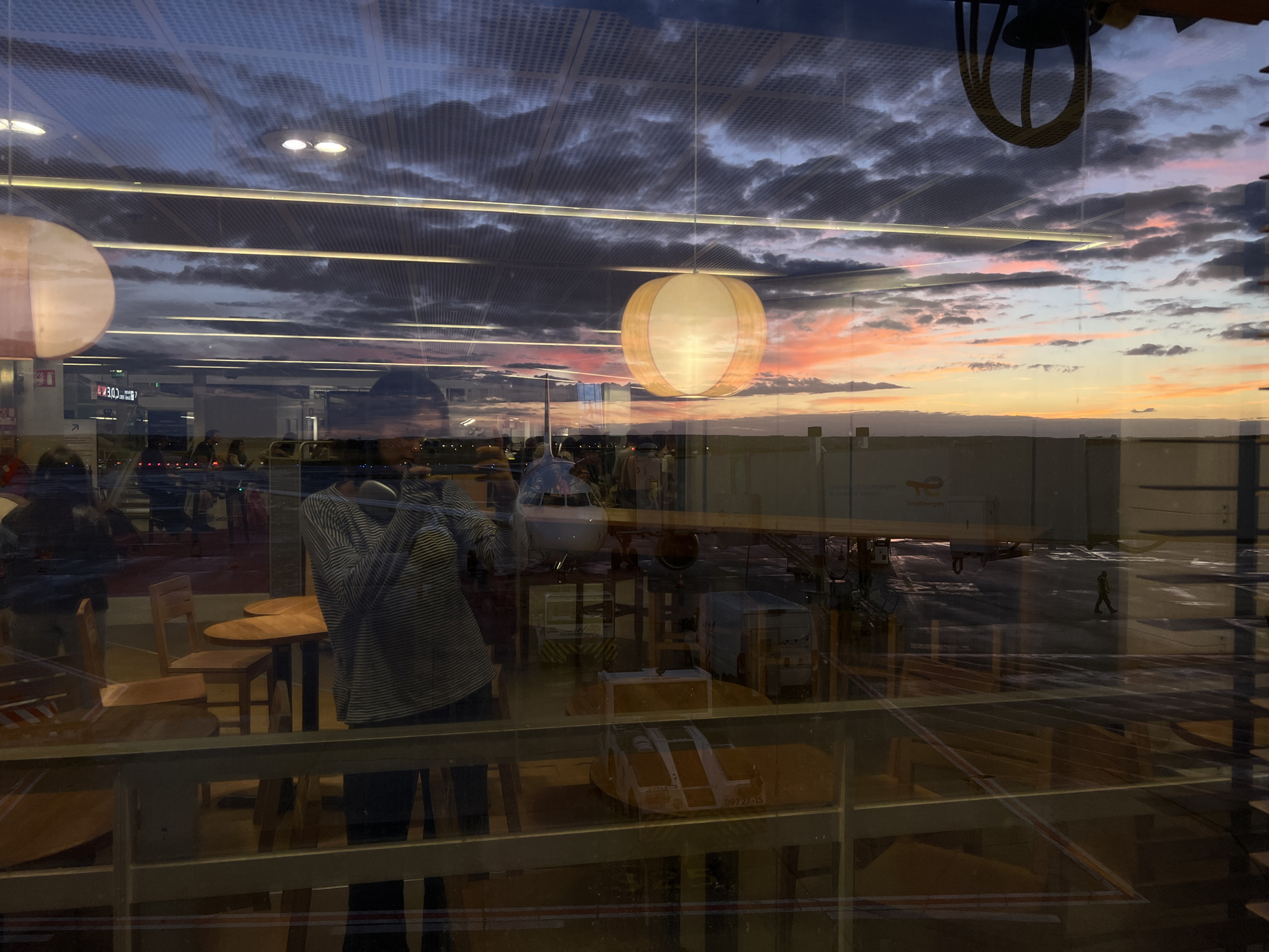 The image shows a reflection on a glass pane, likely at an airport due to the presence of an airplane, jet bridge, and airport vehicles. The reflection displays an interior space with tables and chairs, suggesting a waiting area or restaurant. A person in a striped shirt is visible in the reflection, holding a phone. Outside, a sunset is occurring with dramatic clouds illuminated by orange and pink hues. The location is identified as an airport in Paray-Vieille-Poste, France, which is near Charles de Gaulle Airport. The overall scene captures the transition from day to night with a vibrant sky and the mundane reality of an airport environment.