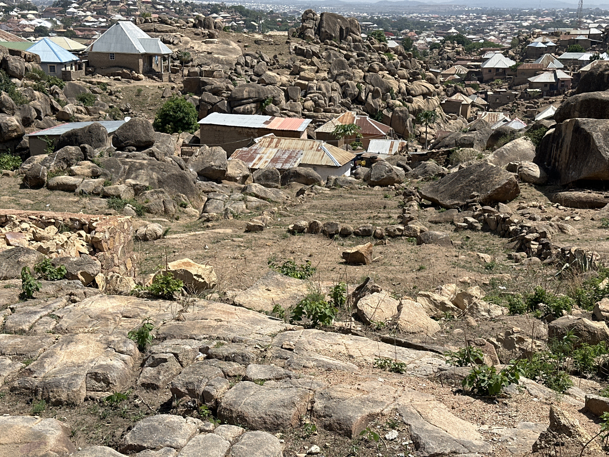 The image depicts a rocky, hilly landscape dotted with buildings, likely located in Jos, Nigeria. The foreground is dominated by large, exposed rocks and dry ground with sparse vegetation. The midground features numerous boulders and houses with corrugated metal roofs, clustered among the rocks. The background shows a denser urban area, extending towards the horizon. The sky is overcast, contributing to the overall muted lighting of the scene. There are no people visible in the frame. The environment is rugged and arid, suggesting a harsh climate.