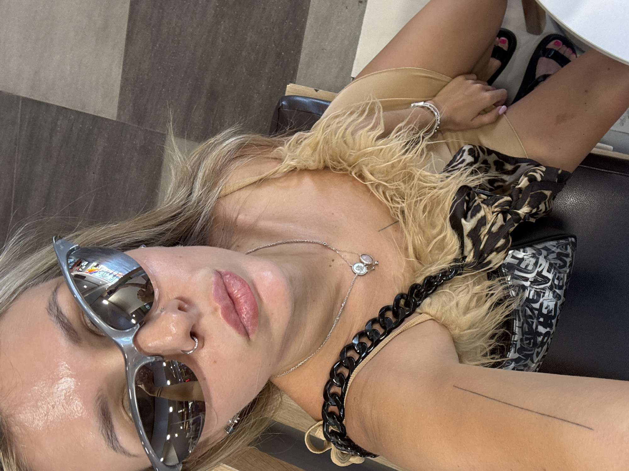 A woman with blonde hair, wearing sunglasses, a tan dress with fluffy trim, and a black chain strap bag, sits in a chair. Her left arm is visible with a straight line tattoo. Her feet are bare, with bright pink nail polish. She is wearing black sandals. The background is a tiled floor in shades of gray and tan. The setting is an indoor location.