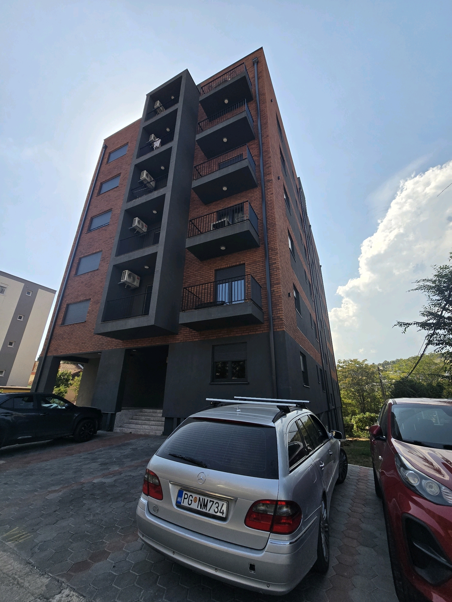 This is a medium shot, low-angle outdoor photograph taken in Podgorica, Montenegro. The primary subject is a modern, multi-story apartment building with a facade of red brick and dark gray accents. The building features several balconies with black railings. Air conditioning units are visible on some of the balconies. In the foreground, a silver station wagon with a Montenegro license plate (PG NM734) is parked, facing away from the camera. To the left of the station wagon, a dark SUV is parked, also facing away. To the right of the station wagon, the front of a red SUV is visible. The ground is paved with hexagonal stone tiles. In the background, lush green trees and a hillside are visible. The sky is bright with a few white clouds, suggesting it is daytime. The overall scene depicts a residential area in Podgorica on a clear day.