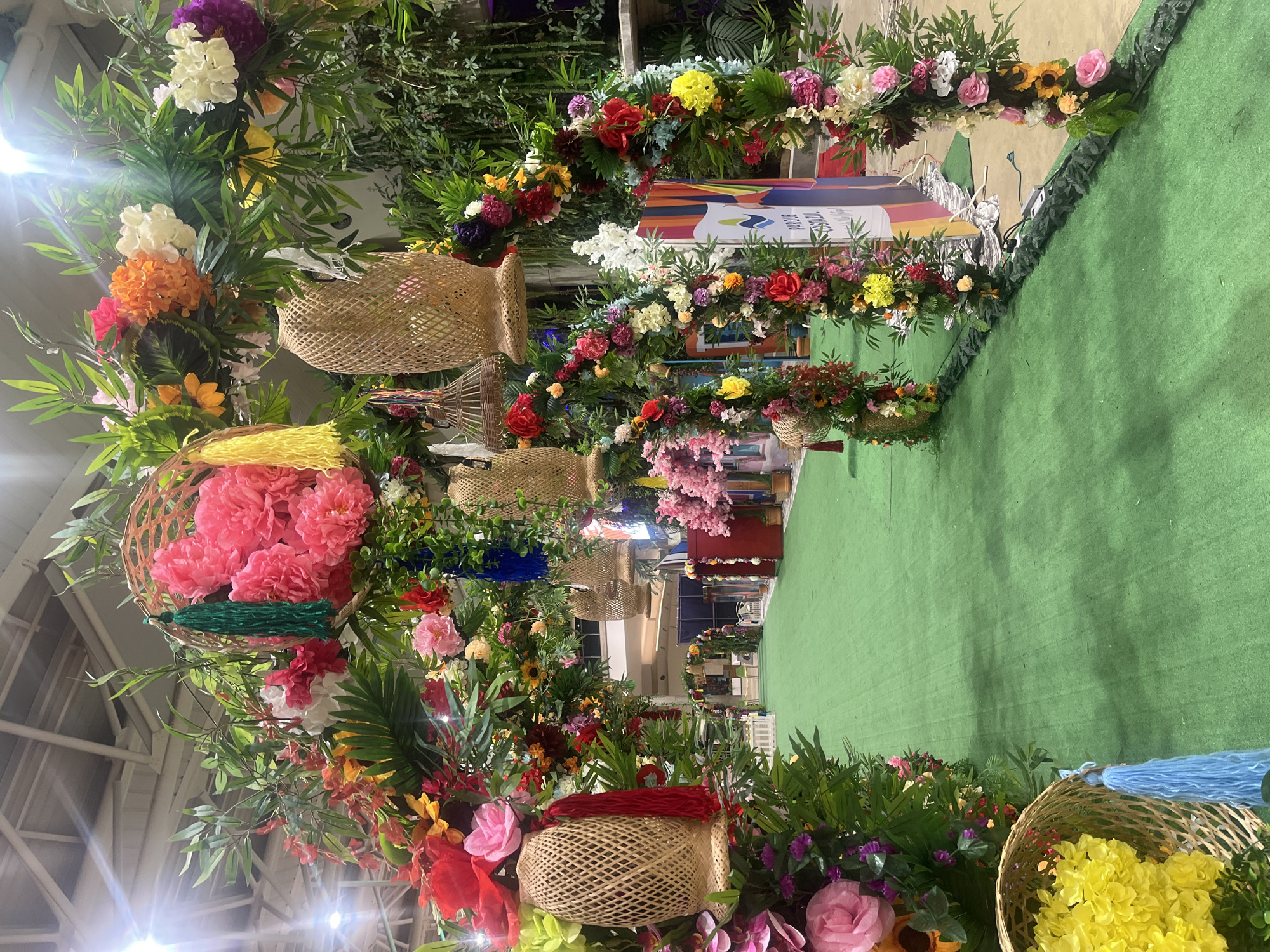 The image captures a vibrant floral display, possibly an archway or decorative installation, in an indoor setting. A wide assortment of flowers in various colors including red, pink, yellow, white and purple are arranged amid green foliage. Interspersed throughout the floral arrangement are woven baskets, some adorned with colorful tassels. The location seems to be a commercial space, with hints of shops or booths visible in the background. A green carpet runs underneath the floral display. A sign featuring a colorful design and the words "Parque Tazajal" is visible. Pampatar, Venezuela is not discernible from the image.