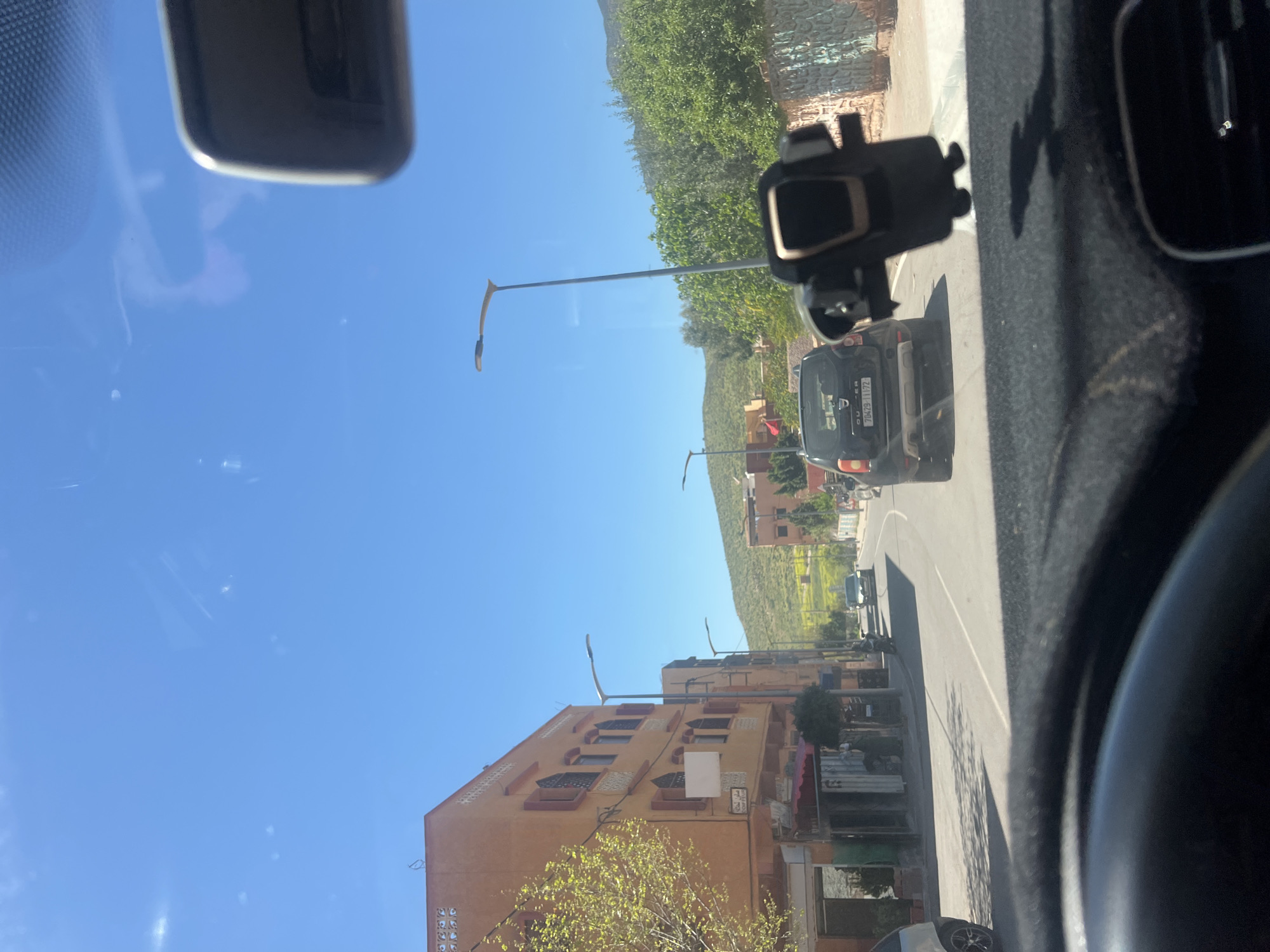 View from inside a vehicle traveling on a street in Ouaouizert, Morocco. The sky is bright blue. Buildings line the street, which curves slightly. The terrain rises in the background. A dark-colored SUV is visible in front of the camera vehicle. Power lines and streetlights run along the street. Trees and vegetation are also visible.