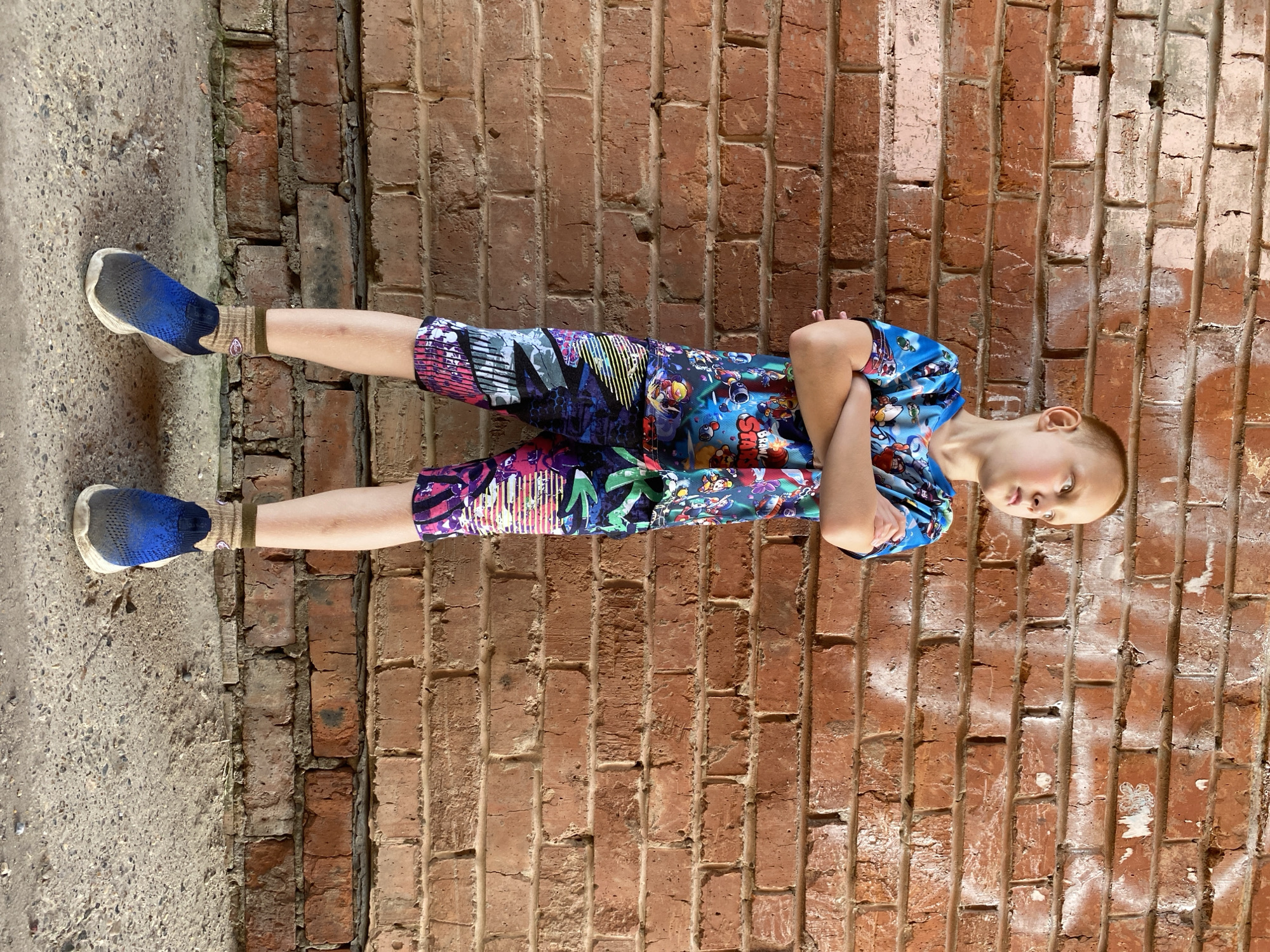 A young Caucasian boy stands with his arms crossed in front of a weathered brick wall. He is wearing a brightly colored t-shirt and shorts with a video game theme and blue athletic shoes. The boy is looking to his left, with a serious expression on his face. The brick wall appears to be part of an outdoor structure, possibly in an urban setting. The ground beneath the boy is concrete. The image is taken from a slightly low angle, emphasizing the boy's stature against the backdrop of the wall. The lighting suggests it is daytime. There are no specific landmarks visible that would indicate the location as Noviy, Russia.