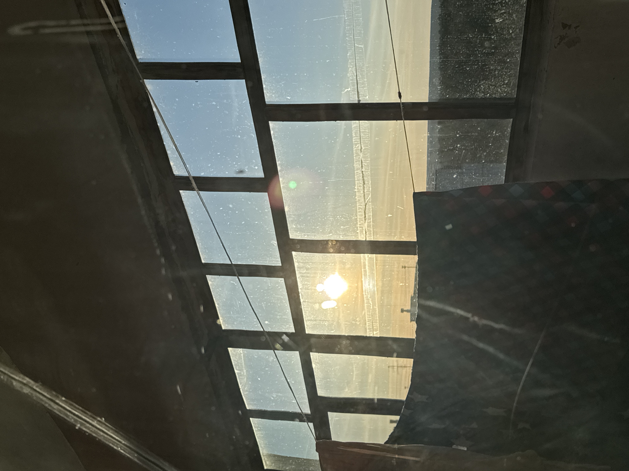 The image presents an interior perspective looking out through a gridded window or skylight, likely in Chaikovsky, Russia. The structure consists of numerous rectangular glass panes set within dark, possibly wooden or metal, frames. The glass appears dusty and streaked, suggesting age or lack of cleaning, which diffuses the incoming light.
A prominent feature is the bright, low-angle sun, directly visible through one of the upper-right panes. This causes significant lens flare, manifesting as a bright, almost square-shaped highlight and a distinct green circle. The intense sunlight bathes the exterior view in a hazy, warm, yellowish glow, making specific outdoor details indistinct, though faint vertical and horizontal elements suggest distant structures or a landscape.
Inside the captured space, dark, undefined surfaces frame the window on the left and bottom. A section of dark, patterned fabric with red, white, and blue diamond-like shapes is visible on the lower right, possibly part of a curtain or blanket. Thin wires or strings run across several panes, with one notable wire stretching diagonally from the top left towards the center. The strong, direct light indicates either early morning or late afternoon. No people or visible text are present in the image.