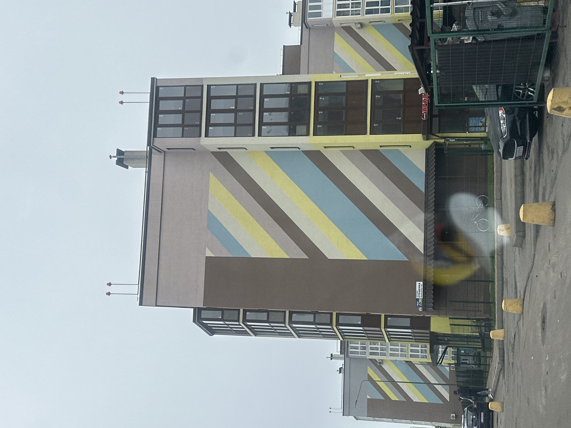 A building is seen in the city of Kyiv, Ukraine. The building has a distinctive stripe pattern. The street sign "75 Стеценка" is visible, indicating the building's location on Стеценка street. The surrounding area consists of pavement and possibly a road. Vehicles are parked nearby, and there is a metal structure present, possibly a dumpster or enclosure. The weather appears overcast.