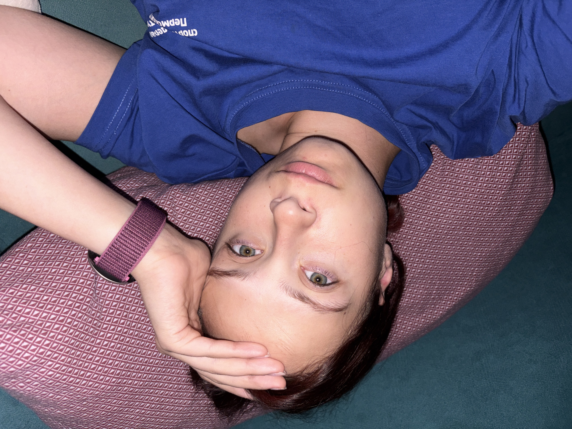 The media file captures a young individual, likely female, lying on a patterned maroon pillow against a teal fabric background, suggesting an indoor setting like a couch or bed. The person is viewed from an overhead perspective, looking directly upwards at the camera with a neutral expression. They are wearing a plain royal blue short-sleeved t-shirt. On their left wrist, a purple or maroon fabric watch band is visible.
The individual has dark brown or reddish-brown hair and green or hazel eyes. One hand is casually resting on their forehead, partially obscuring the hairline. The pose implies relaxation or resting.
White text is printed across the chest of the blue t-shirt. From the viewer's perspective, this text appears upside down. When correctly oriented, the text reads "СПОРТ Пермь" (SPORT Perm). The time of day is not discernible from the indoor lighting. The location context for this media file is Прикамский, Russia.