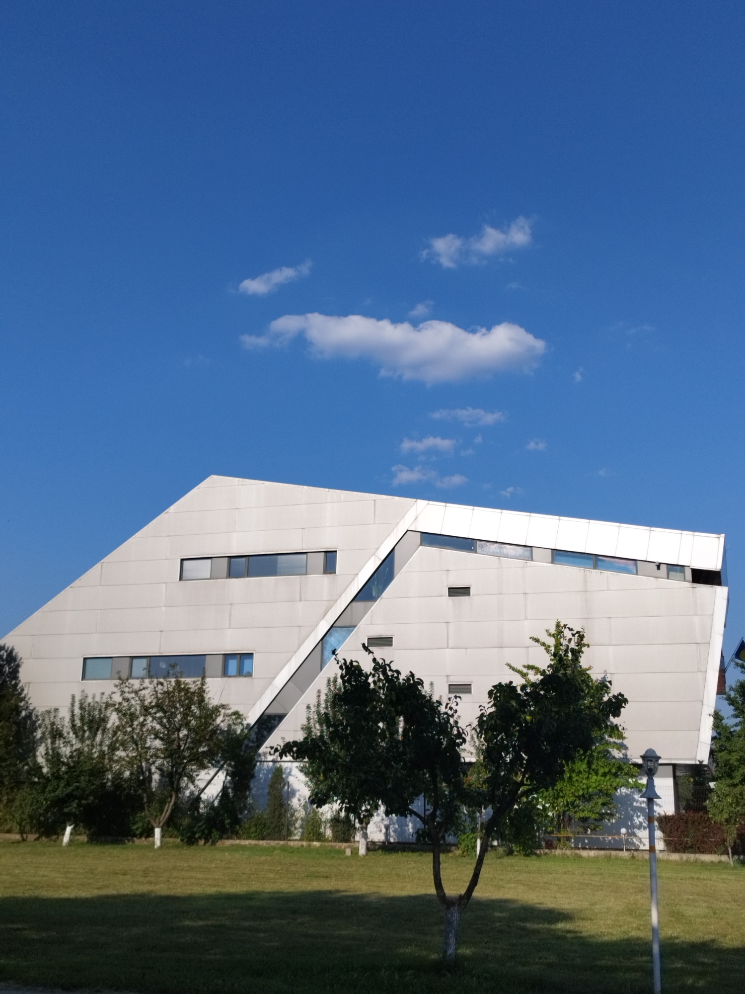 This image depicts a modern, angular building with a white, metallic facade, situated in a grassy area with several trees. The building's architecture is characterized by sharp lines and geometric shapes, with long, horizontal windows. A diagonal section of glass cuts across the facade, reflecting the blue sky. The sky is clear with a few scattered white clouds. The foreground consists of a well-maintained lawn, and several trees are visible in front of and around the building. The overall scene suggests an outdoor environment, possibly in a park or a campus setting. There are no people visible in the image. The building's unique design suggests it could be a contemporary public building, such as a library, museum, or administrative center. While the exact location in Bucharest, Romania is not specified, the architectural style is consistent with modern developments in urban areas.
