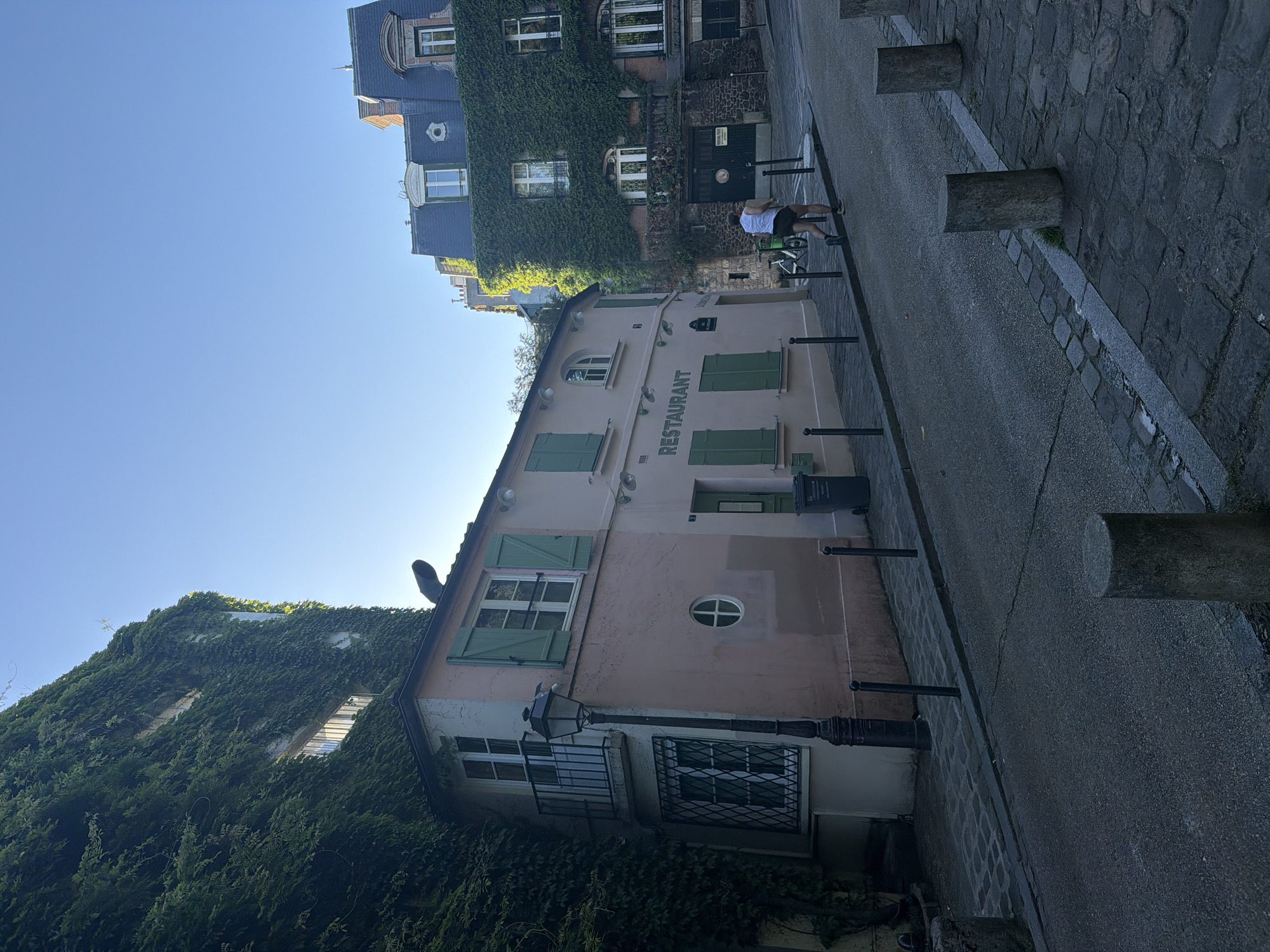 A street-level, daytime image of a street with buildings in Montmartre, Paris, France. One building houses a restaurant and it is peach-colored with green shutters. The word "RESTAURANT" is painted vertically on the building's facade. Ivy grows on the buildings. A person wearing a white top and dark shorts is seen riding a bicycle on the street. The street is paved with cobblestones and asphalt. Bollards line the street. The sky is blue.