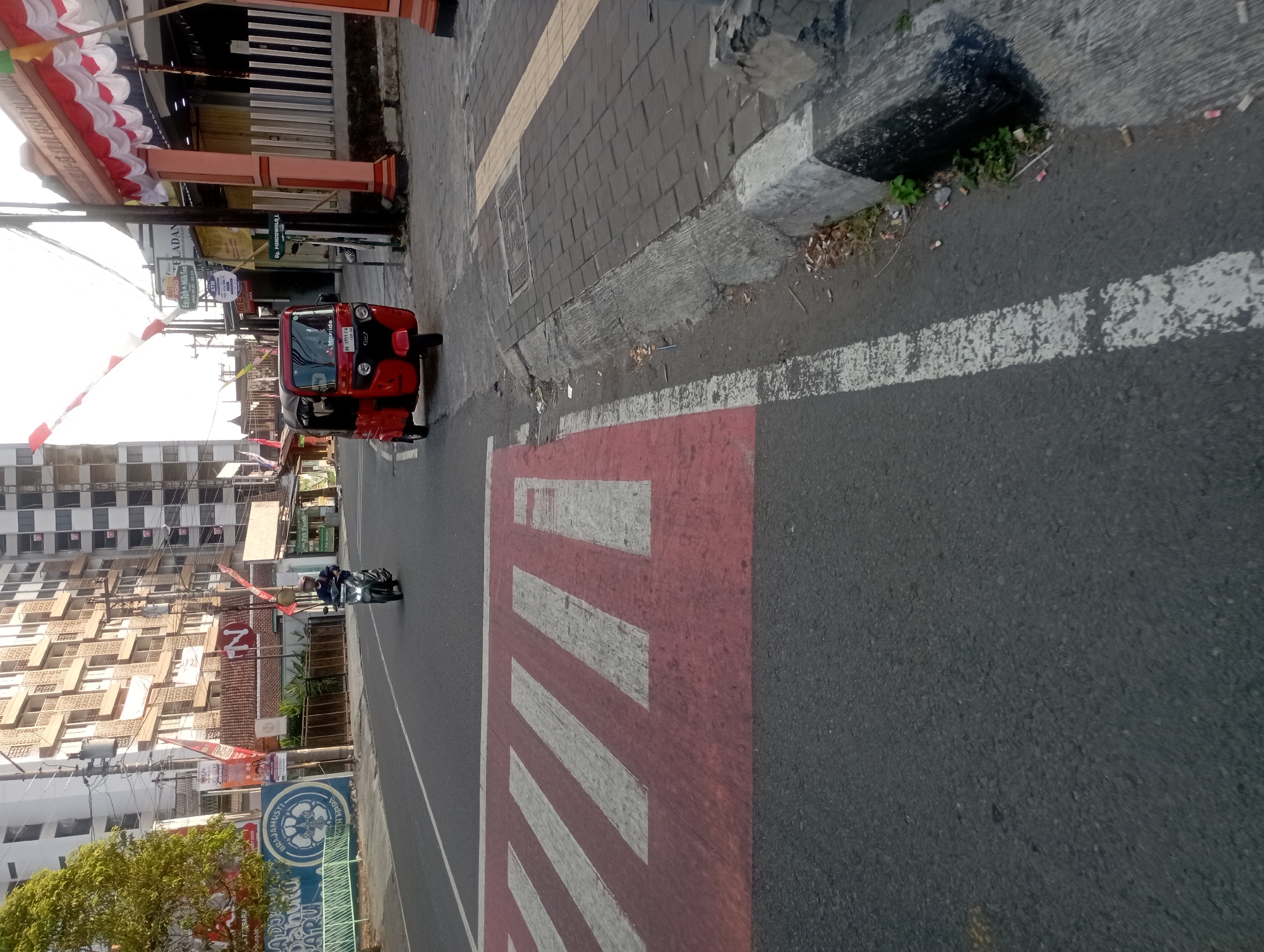 This is a street-level, eye-level, medium shot of a street in Yogyakarta, Indonesia. In the foreground, a red and black tuk-tuk is stopped in the middle of the street, its license plate visible as AB 3519 RW. To the left, a pedestrian crossing with red and white stripes is painted on the asphalt. A person on a scooter, wearing a helmet, is riding past the tuk-tuk. In the background, a tall, modern building with numerous windows dominates the skyline, with a large tree on the left side of the image. The street appears to be relatively empty, with minimal traffic. The overall scene suggests a typical urban environment in Yogyakarta.
