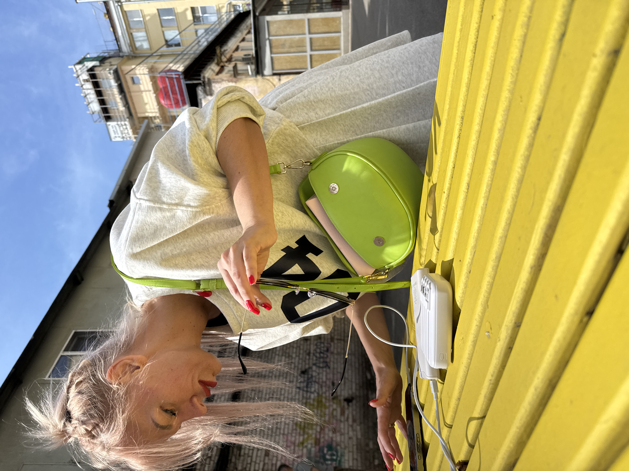 In Saint Petersburg, Russia, a woman with pink-tinted hair pulled into a bun and braid, wearing a grey crewneck sweatshirt and a pleated skirt, is leaning over a bright yellow picnic table. She is holding a pair of sunglasses in her right hand, examining them. On the table in front of her are a smartphone, a white power bank, and a white charging cable. To her right, a lime green crossbody bag with a tan interior is resting on the table. The background shows a gritty urban environment with brick walls covered in graffiti and multi-story buildings under a clear blue sky.