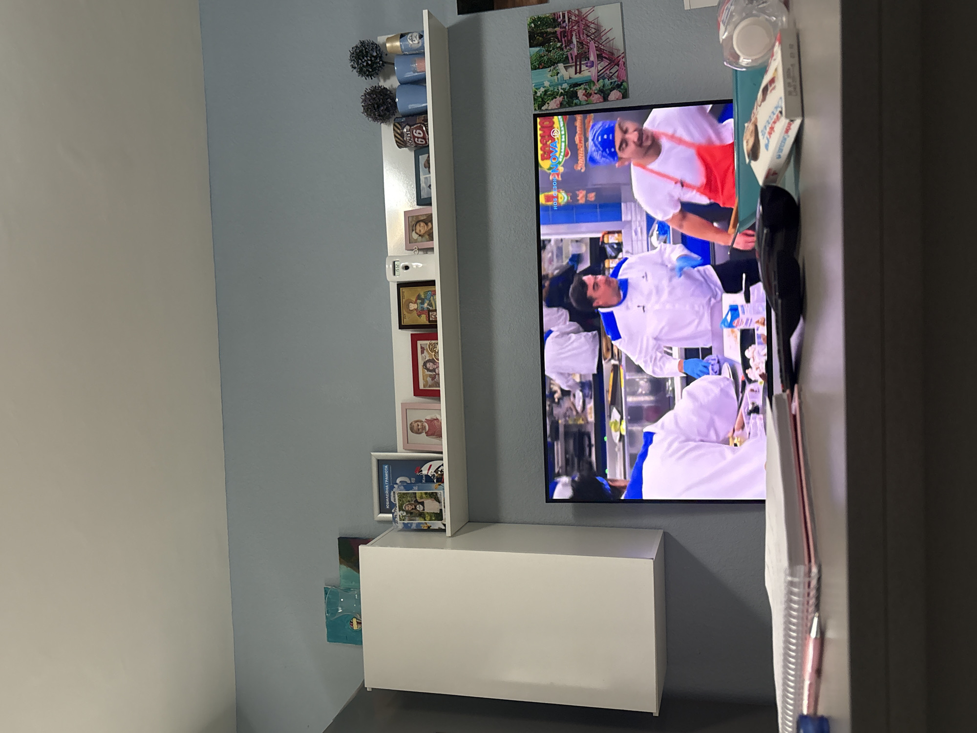This media file captures an indoor scene in Sofia, Bulgaria, likely a living room or entertainment area, featuring a television and decorative shelving. The image is taken from a slightly upward and rotated perspective, showing two distinct wall colors: a light blue-grey behind the main display area and a lighter beige-white on the adjacent wall.
The central subject is a flat-screen television, mounted on the light blue-grey wall, displaying a cooking show. On the screen, several male chefs are visible. One chef in the upper portion wears a white uniform with a red apron and a blue bandana. Another chef, prominent in the lower half of the screen, wears a white uniform with blue accents and blue gloves, actively engaged in food preparation. Visible text overlays on the TV screen include "HELPS NOVO" and "ROB," alongside other fragmented text in a non-English language.
To the left of the television, a white floating shelf is adorned with various objects. These include two small, dark, spherical decorative plants, two light blue cylindrical containers or vases, and a miniature "Route 66" sign. Below these, multiple framed photos are displayed, one showing a close-up of a person, and another larger frame containing text that appears to be a "КОМИСИОННА ГРАМОТА" (Commissioned Diploma/Certificate in Bulgarian). A digital clock on the shelf reads "15:52."
Below the television, a white rectangular cabinet or storage unit is mounted on the wall. To the right of the TV, part of a table or desk is visible, holding a clear plastic bottle and a box of "Kinder Choko-Bons." A black object, possibly a remote control or a phone, and a pen are also present on this surface. Above the television, a piece of wall art depicting a scene with pink and blue elements is mounted. The overall lighting suggests it is daytime indoors.