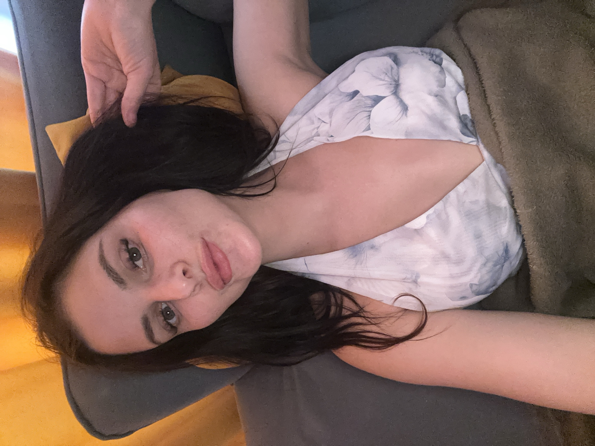 A woman with dark hair and fair skin is lying on a sofa, wrapped in a blanket. She is wearing a white halter top with a floral pattern. Her right hand is raised to her hair, and her left arm is resting on her side. The sofa is dark gray, and a yellow pillow is visible behind her. The background is a blurry yellow curtain. The scene appears to be indoors.