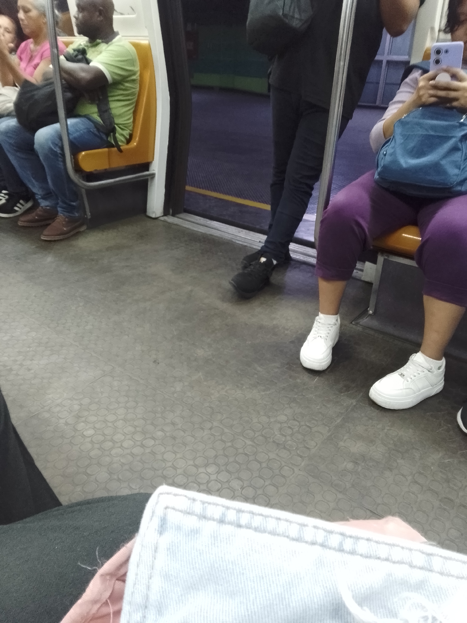 The photograph shows a subway car interior. Four people are partially visible: One woman sits in a pink shirt and a man in a green shirt and jeans sit facing forward. A man in black pants and a jacket stands in the aisle, while a woman sits in purple pants and white shoes using her cell phone. A blue purse sits in her lap. The subway car has yellow seats with metal frames. The subway car floor has a patterned surface. The subway car is located in Caracas, Venezuela.