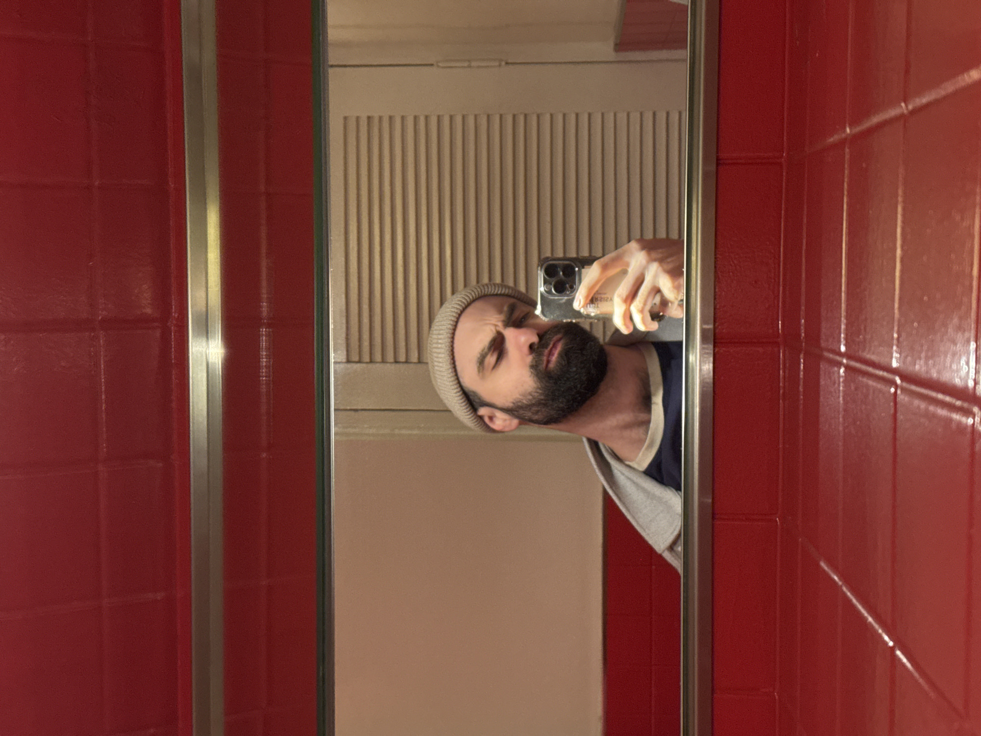 A man with a beard and a beige beanie is taking a selfie in a mirror. He is holding a smartphone in his right hand. The mirror is surrounded by a red tiled wall with silver trim. Behind the man's reflection, there is a white louvered door or vent.
There is no location information provided to identify the city as Yerevan, Armenia. The image does not offer enough environmental information to confirm this detail.