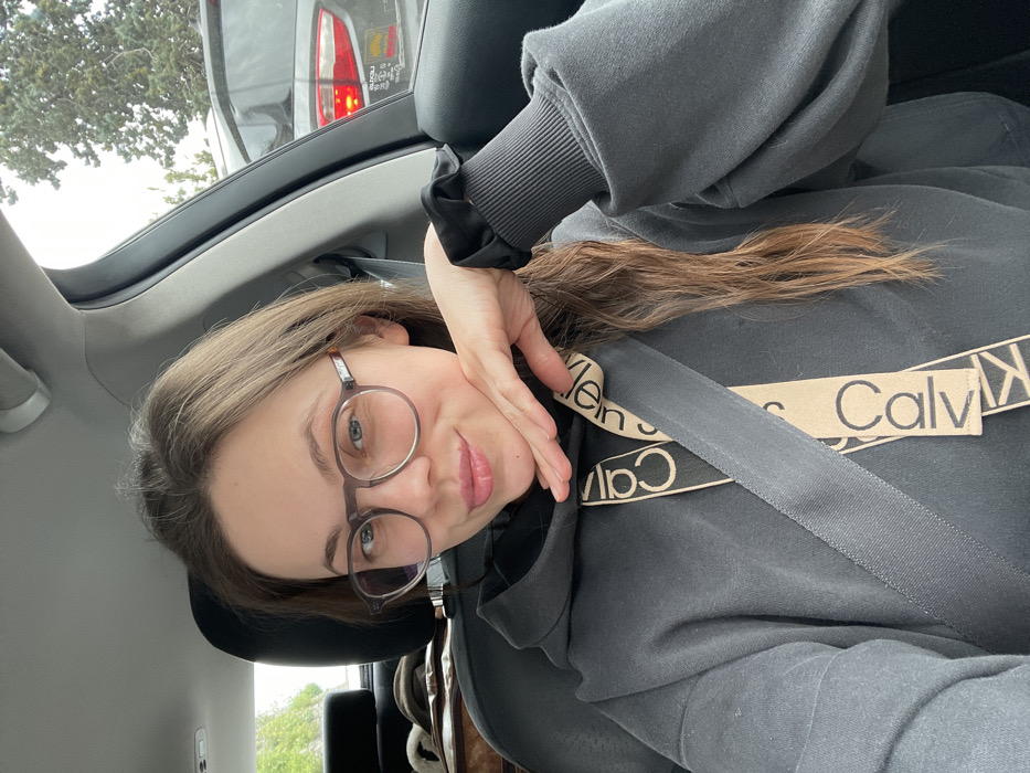 The image displays a young female seated in the passenger seat of a vehicle, identified as being in Ravello, Italy. The female subject has light brown hair, parted on the side, extending over her left shoulder. She wears round, translucent-framed eyeglasses and a dark gray hooded sweatshirt featuring ribbed cuffs. A beige-colored strap, marked with "Calvin Klein" branding in black text, is visible around her neck area, positioned beneath a dark gray seatbelt that diagonally crosses her chest. Her left hand is raised, with fingers resting under her chin, and a black scrunchie is visible on her left wrist. She is looking directly at the camera with a slight smile. The car's interior features dark upholstery and a light gray headliner. Through the passenger window, an overcast sky, green tree foliage, and the rear of a dark gray SUV are visible. The SUV's red taillight is illuminated, and a sticker is present on its rear window. Objects, possibly fabric or a hat, are discernible on a surface behind the subject's seat.
