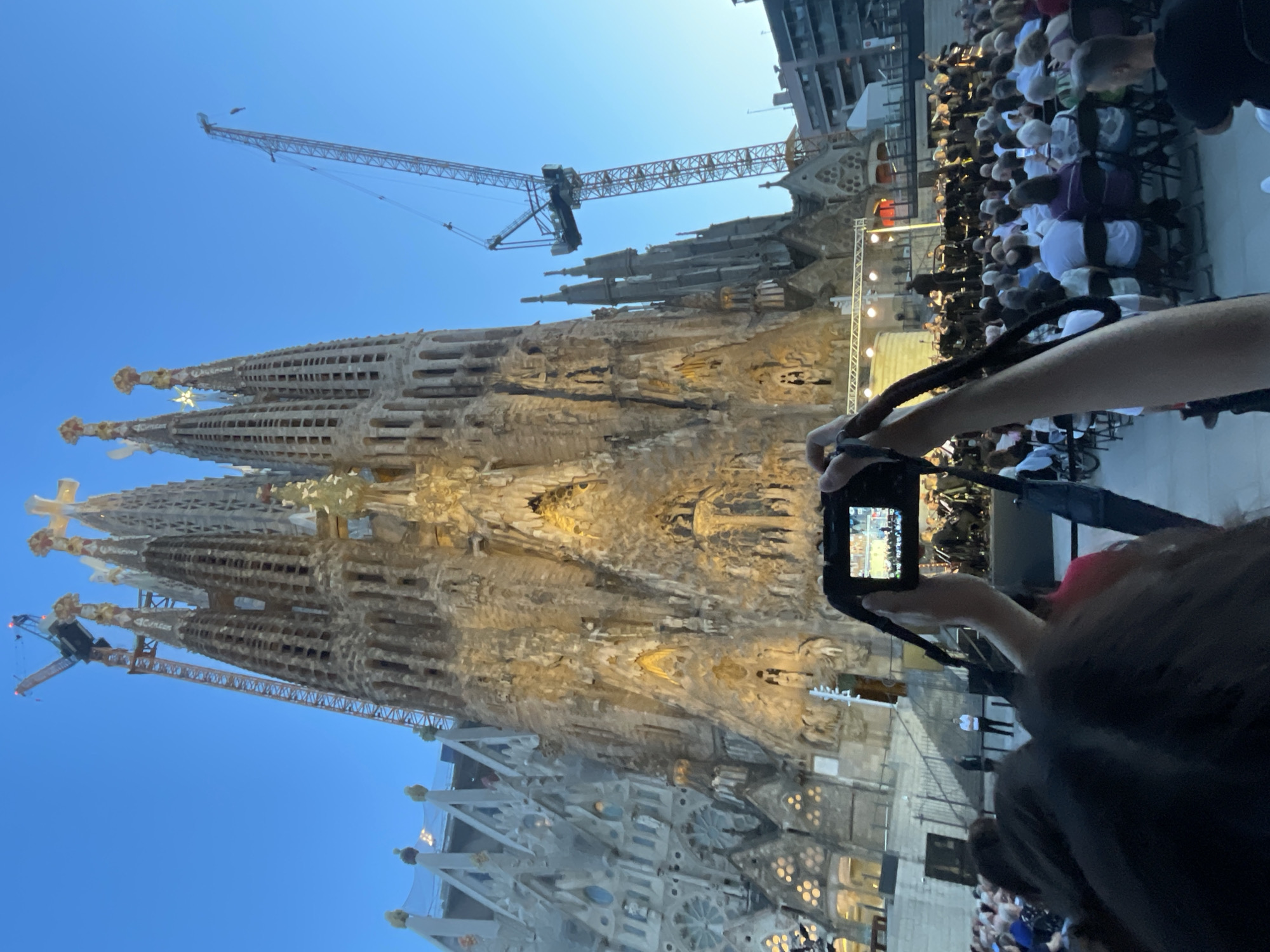 The image depicts the Sagrada Familia in Barcelona, Spain, a renowned basilica under ongoing construction. In the foreground, a person is holding a digital camera, capturing a photograph of a live performance taking place on a stage in front of the basilica. A seated audience is visible, facing the stage. The basilica itself is elaborately decorated with intricate carvings, and scaffolding and construction cranes are present, indicating ongoing work. The sky is clear and blue, suggesting it is daytime. The scene captures a blend of cultural event and architectural marvel.