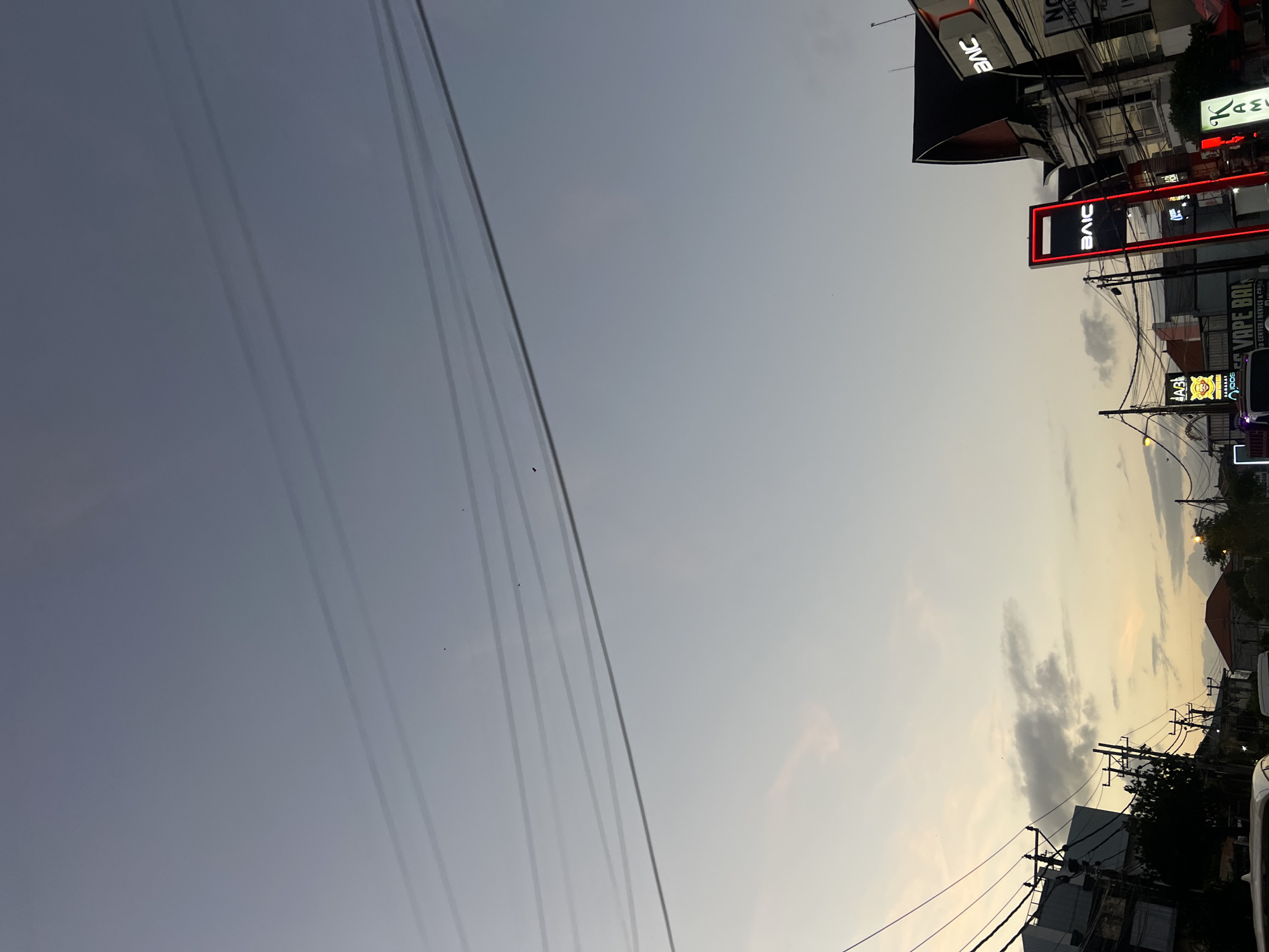 This image depicts a street scene at dusk in Denpasar, Indonesia, with a focus on the sky and the surrounding urban environment. The sky is a pale gradient of gray and light yellow, with thin, wispy clouds visible near the horizon. Several power lines stretch across the frame, casting faint shadows on the sky.
Below the sky, the city emerges with a mix of buildings and commercial signage. A prominent neon sign reading "BAIC" in red and white is centrally located, illuminated against the dimming light. Adjacent to it, another sign also displays "BAIC". To the right, a building with a distinctive dark, pitched roof is visible. Further to the left, other signs are partially obscured, including one with text that appears to be "VAPE BALI". Utility poles with numerous wires are scattered throughout the lower portion of the image, typical of urban infrastructure. A few small birds are seen in the sky. The scene suggests a busy commercial street in Denpasar transitioning from day to night.
