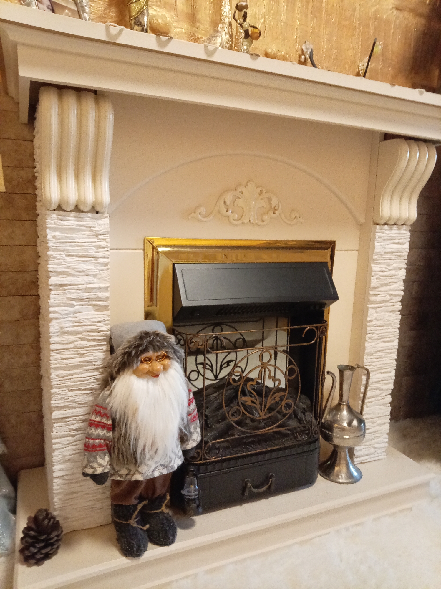 A gnome doll with a white beard and glasses stands to the left of an electric fireplace. The gnome is wearing a patterned sweater and brown pants. To the right of the fireplace is a silver pitcher. The fireplace has a gold frame and a black insert with decorative metalwork. The fireplace is surrounded by decorative pillars and a mantelpiece. The floor in front of the fireplace is covered in white, fluffy material. A pine cone is on the left side of the fireplace. The scene appears to be indoors, likely in a home in Yessentuki, Russia, possibly decorated for the holidays.