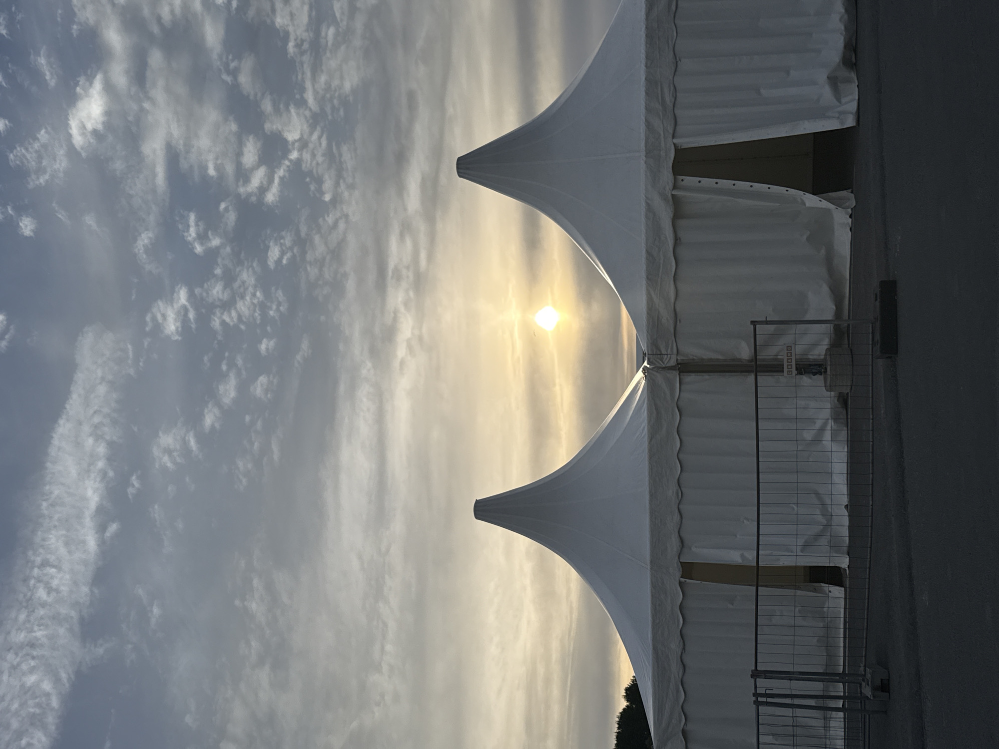 The image shows two white, peaked tents set up side-by-side outdoors. The sky above is partly cloudy with the sun visible, casting a soft glow through the clouds. In front of the tents, a temporary metal fence with a concrete base is installed. The tents appear to be modular structures, likely for an event, with the entrances partially open, revealing darkness within. The ground in front of the fence is paved. The scene is consistent with an outdoor event space in Tallinn, Estonia, possibly during the early morning or late evening, given the lighting conditions.