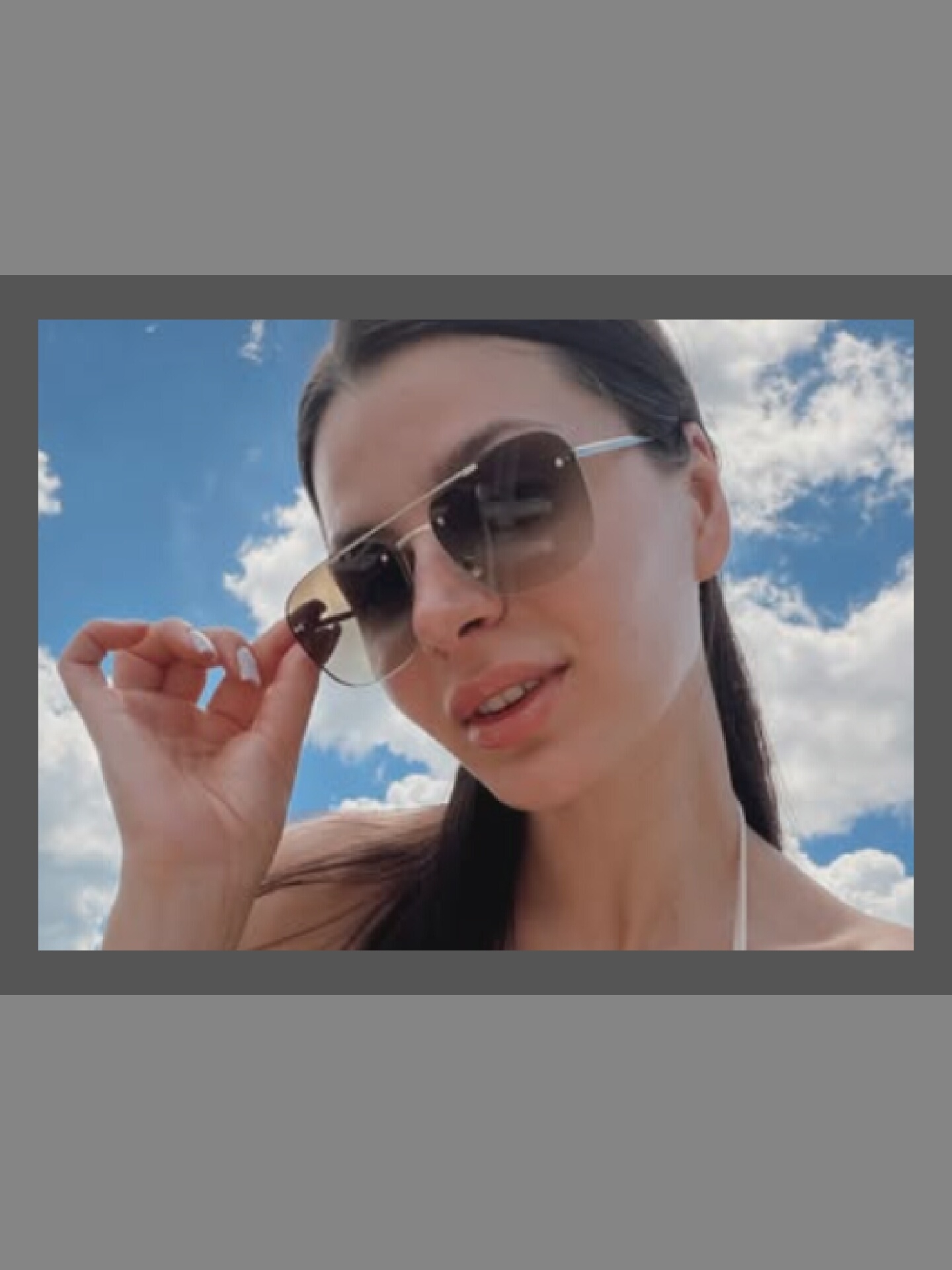 A close-up, low-angle, outdoor portrait shows a young woman with long dark hair, wearing large, rimless sunglasses with gradient lenses. She is looking towards the viewer and has her left hand raised, with her index finger touching the edge of the sunglasses. Her mouth is slightly open, revealing her teeth and lips. She appears to be wearing a white bikini top, the strap visible over her shoulder. The background is a bright blue sky with white, fluffy clouds, suggesting a sunny day. The image is cropped with a gray border on the top and bottom.