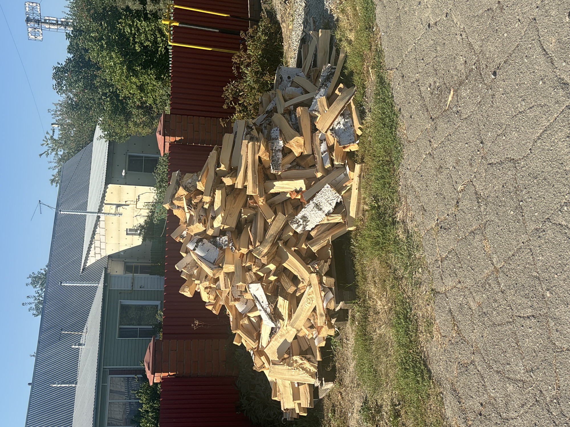 The image captures an outdoor scene in Ob, Russia, on a clear, sunny day. In the foreground, a substantial pile of chopped firewood, predominantly birch with its characteristic white bark, is stacked on wooden pallets. The firewood varies in size and shape, suggesting it has been prepared for burning. Behind the firewood, a red corrugated metal fence runs horizontally across the frame, punctuated by brick pillars. Beyond the fence, two houses are visible. The house on the left has a green siding and a gray metal roof, while the house on the right is constructed with light-colored blocks and also has a gray metal roof. Trees and foliage are present between and behind the houses. The ground in the immediate vicinity of the firewood is covered in dry grass and gravel, while a paved pathway made of interlocking gray stones occupies the lower portion of the image. The sky above is a clear, light blue, indicating fair weather. No individuals are present in the scene.