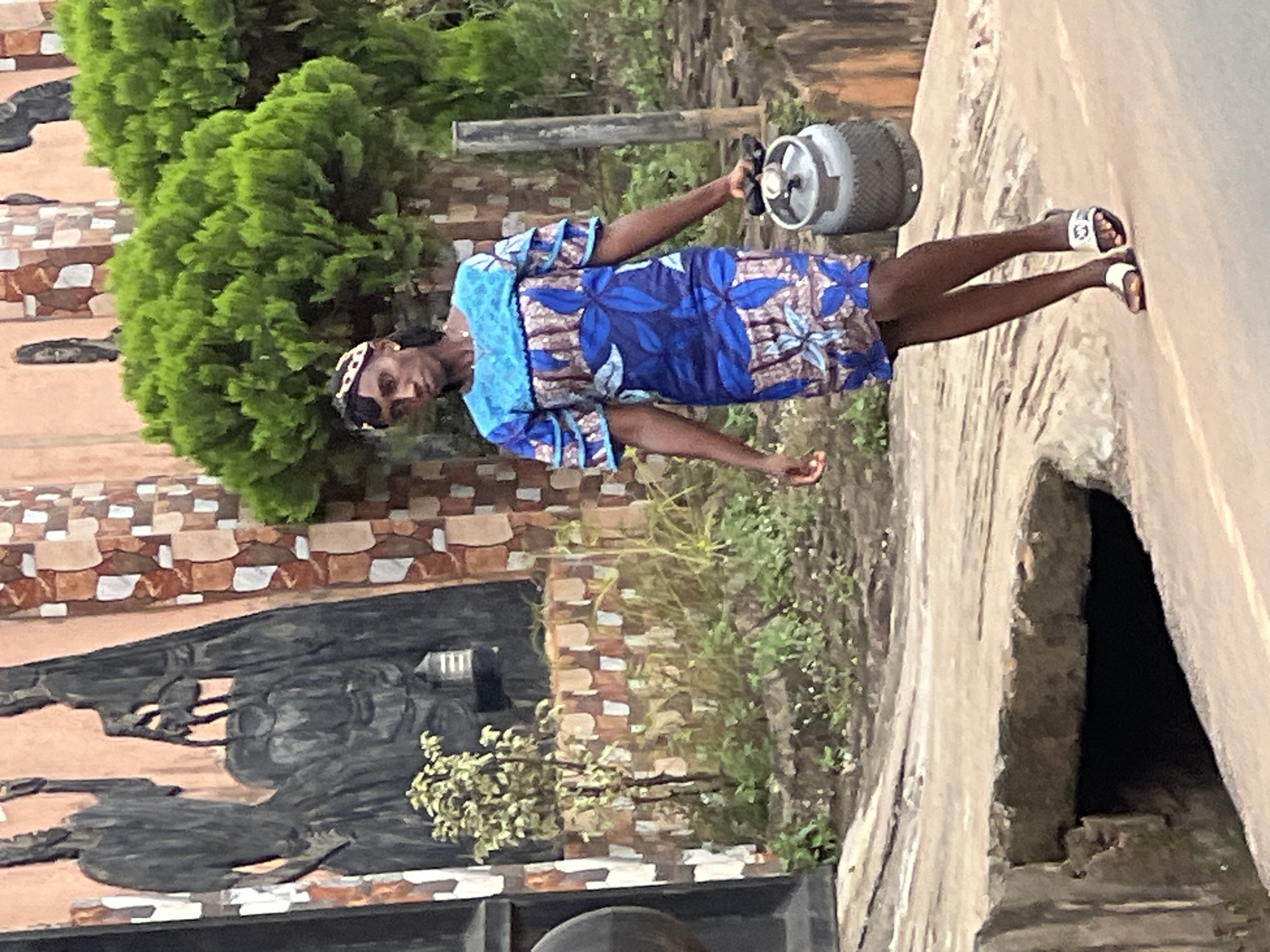 This media file shows a person standing on a paved surface, possibly a roadside, in Nkpor, Nigeria. The person is wearing a blue and white patterned dress and white sandals. They are holding a grey gas cylinder in their right hand and appear to be looking towards the camera. To the left of the person is a green, bushy plant and a building with a decorative facade made of brick and white and brown tiles. The building has a dark, possibly metal, entrance structure. The overall scene suggests an outdoor environment during daylight, with a cloudy or overcast sky, as indicated by the diffused lighting. The presence of the gas cylinder suggests the person is engaged in an activity related to obtaining or transporting cooking gas, a common occurrence in many Nigerian households. No visible text is present in the image.