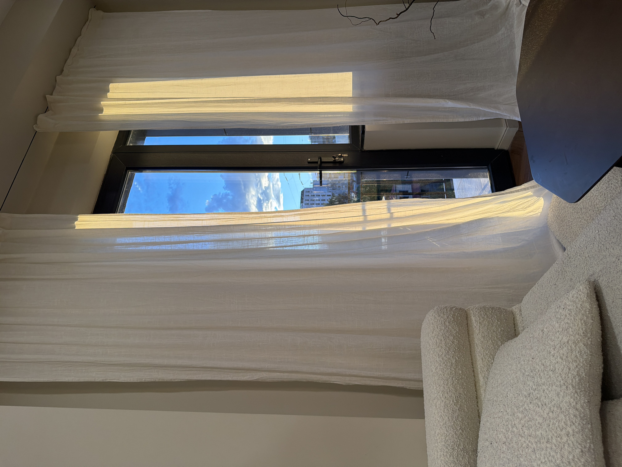 The image shows a window with white curtains, partially open, revealing a view of Yerevan, Armenia. The window has a black frame and is equipped with a latch. Outside the window, one can see buildings, including what appear to be apartments, and a cloudy sky. Inside the room, there is a white, textured armchair and a dark-colored table partially visible in the corner of the room. The overall environment seems to be a well-lit interior space.