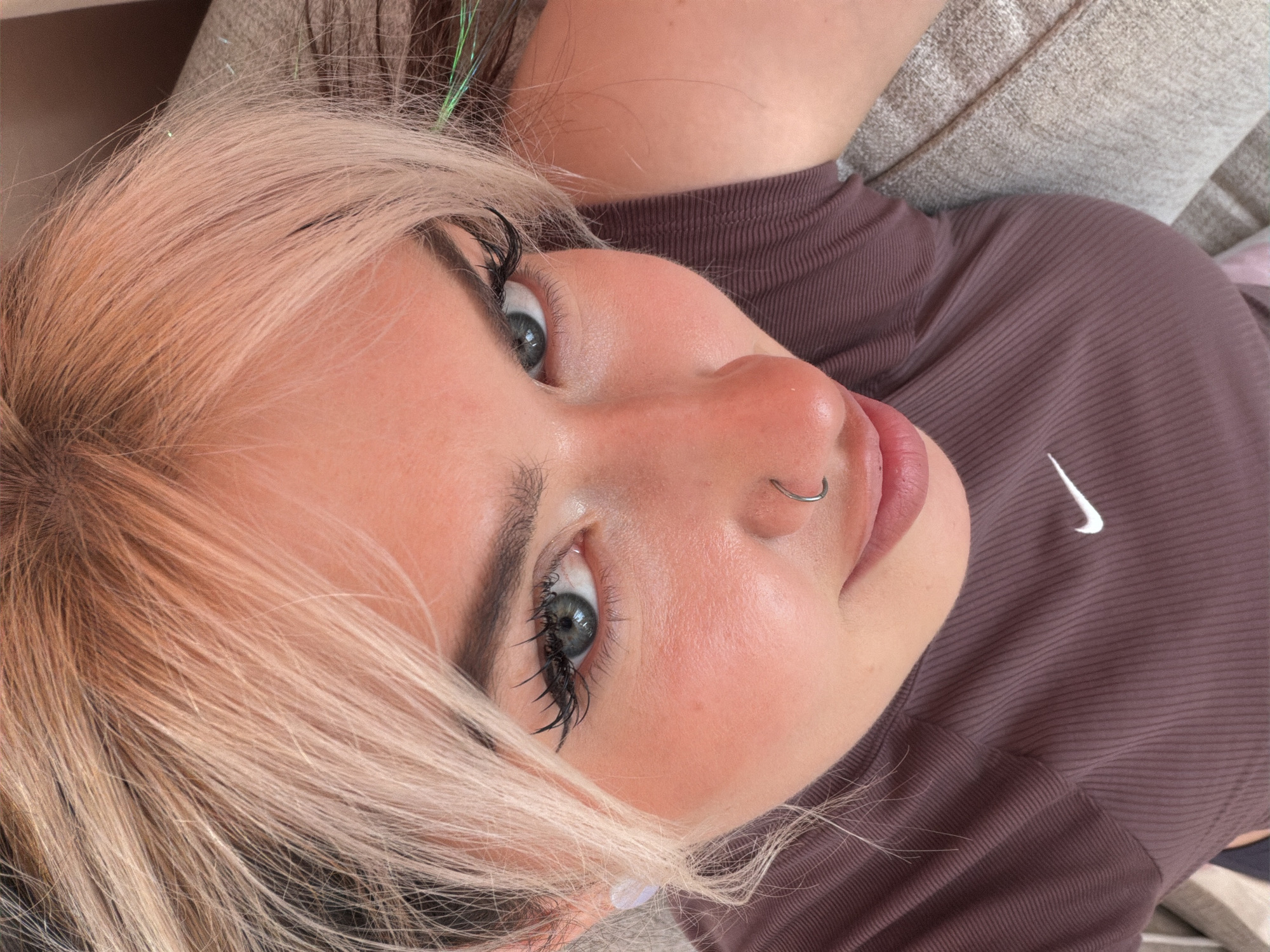 A person lies on a beige couch. The person has light, pinkish-blonde hair with some green tinsel strands visible. The person's face is prominent, showing green eyes, full eyelashes coated with mascara, a nose ring, and pink lips. The person is wearing a brown, ribbed shirt with a white Nike swoosh on the upper right side. The couch appears to be in a domestic environment; no specific details suggest the city of Budva, Montenegro.