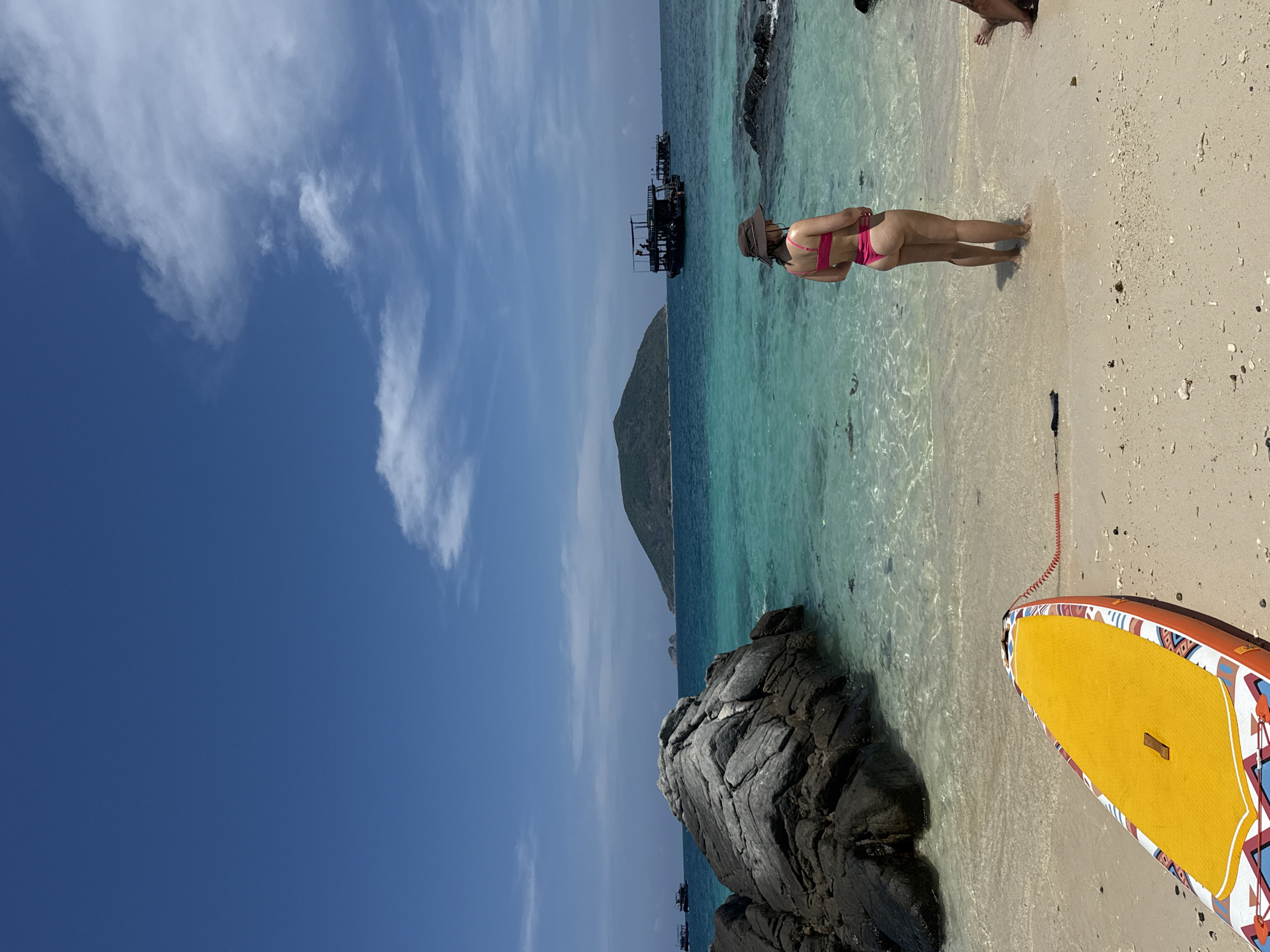 The image shows a woman in a pink bikini standing in shallow, clear water at a beach. She is wearing a hat. A paddleboard rests on the sand next to her. Behind her, there is a rocky area, a small island with a peak, and what appear to be docked boats or platforms in the distance. The sky is blue with scattered clouds. The location is Nha Trang Ward, Vietnam, indicated by the presence of a small island that closely resembles Hon Mun Island located in Nha Trang Bay.