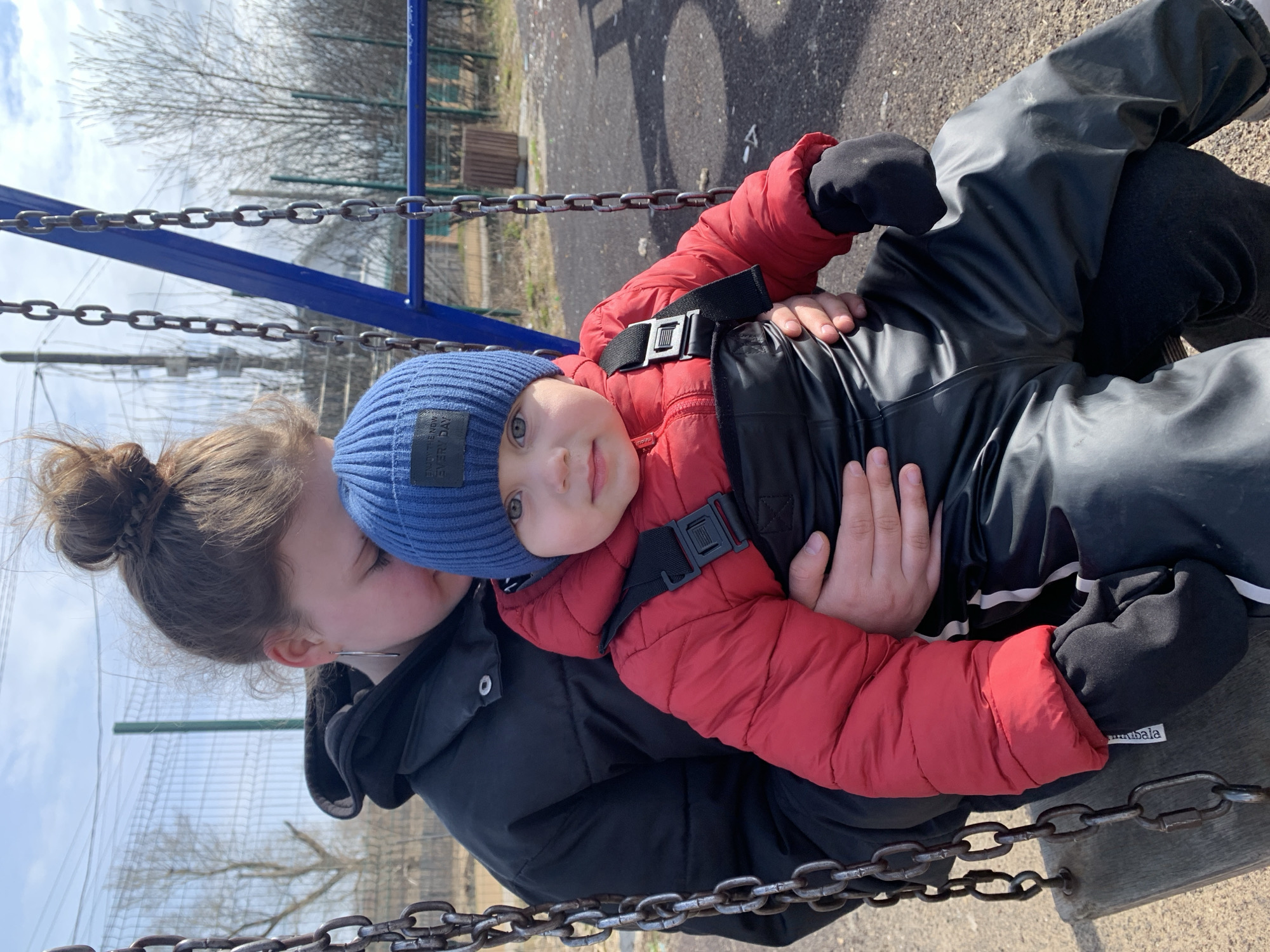 The image captures an adult holding a young child in an outdoor setting, likely a playground in Фоки, Russia. The subjects are positioned horizontally, as if lying down or leaning back together, possibly on a swing or bench, under a bright, partially cloudy sky during daytime. The cool weather, suggestive of late winter or early spring, is indicated by the bare trees in the background and the warm attire worn by both individuals.
The adult, whose face is partially obscured by the child's head, has light brown hair tied in a messy bun and wears a black padded jacket. An earring is visible on the adult's ear.
The child, appearing to be a toddler, looks directly at the camera with a calm expression. They have light eyes and are dressed warmly in a blue knit beanie, a vibrant red padded jacket, and black waterproof overalls with suspenders. A black strap with a buckle is visible across the child's chest. The blue beanie features a label with the text "ENJOY THE NOW EVERY DAY." A white label with "Imubala" is visible on the child's left glove or mitten.
The background shows prominent metal chains from a swing set, along with a blue metal support structure. Beyond this, bare deciduous trees, a green mesh fence, and a dark paved surface complete the outdoor scene.