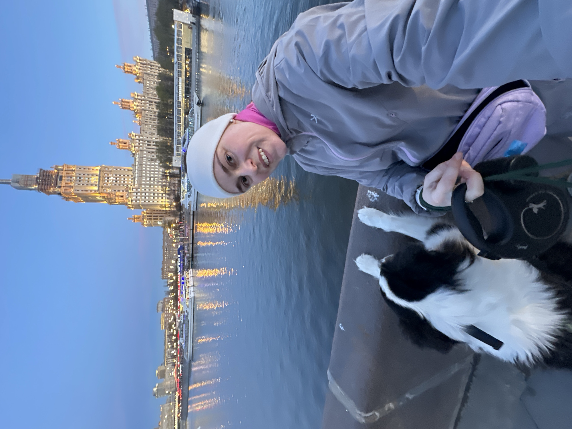 The image captures a woman and her dog outdoors during dusk or early evening, positioned along a river in Moscow, Russia. The photograph appears to be a selfie taken by the woman.
In the foreground, a smiling woman, wearing a white beanie, a pink top, and a grey hooded jacket, looks directly at the camera. She holds a black retractable dog leash in her right hand, and a lilac-colored fanny pack is visible on her left hip. Partially visible in the bottom right corner is a black and white dog with fluffy fur, appearing to look down from a dark grey concrete barrier or embankment. Its white paws are clearly seen.
The background showcases a wide river, likely the Moskva River, reflecting the numerous golden and colorful lights of the city. Along the far bank, a prominent, multi-spired building, characteristic of Moscow's "Seven Sisters" Stalinist skyscrapers, stands brightly illuminated against the deep blue twilight sky. Other buildings further along the riverbank are also lit, and several illuminated boats or ships can be seen on the water. The sky is a clear, dark blue, indicating the transition from twilight to night. The woman's smile suggests a happy and relaxed mood, enjoying the scenic view with her dog. Faint, unidentifiable logos are present on the woman's jacket and the dog's leash.