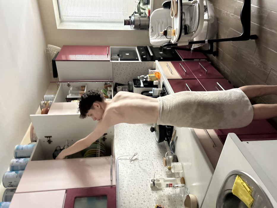 An interior shot depicts a domestic kitchen located in Noviy, Russia. A young adult male, shirtless and wrapped in a light beige towel around his waist, stands facing away from the viewer, his right arm extended into an open upper kitchen cabinet. His dark, curly hair is styled short.
The kitchen features a light-colored countertop with a floral-patterned backsplash. On the countertop, from left to right, are a stainless steel sink with a yellow microfiber cloth, a glass bottle, a decorative glass jar, and a black cooking pot on a stovetop. Various small bottles and containers are also visible. The lower cabinets and drawers are a maroon color with silver handles.
Upper cabinets are a mix of light pink/maroon and white; one white door is open, revealing dishes on a drying rack. Above the upper cabinets, several containers, including what appears to be baby formula cans, are stored. To the right, an integrated oven is visible beneath a black-faced appliance, possibly a microwave or coffee machine, set into a white cubby. A silver juicer or blender rests on the far right section of the countertop, near a window fitted with horizontal blinds. A white high chair with a tray, containing a baby bottle and a divided plate, is positioned near the window. The floor is light-colored wood or laminate.