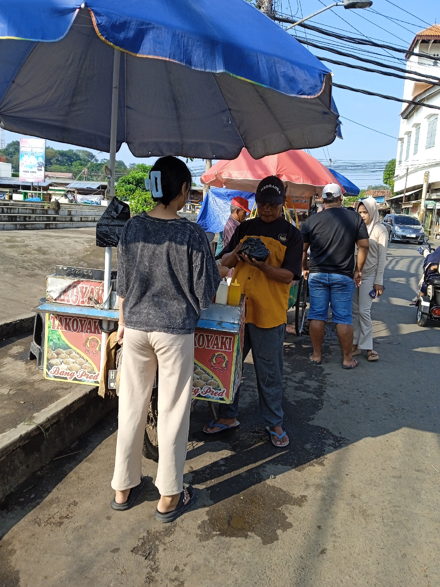 In Kaliwungu, Indonesia, a woman with a blue hair clip is standing at a food cart labeled "TAKOYAKI Bang Pred," purchasing something from a vendor. The vendor, wearing a "Nebraska" cap and an orange and black shirt, is holding a dark object in his hands, possibly food in a bag. They are standing under a large blue umbrella that shades the food cart.
Behind them, there are other customers waiting: a man in a black shirt and denim shorts, and a woman wearing a hijab. A gray car is parked on the side of the road and a motorcycle is parked behind the woman in hijab.
The setting appears to be a street-side vendor area. In the background, there is a building and trees. The sky is visible and it appears to be a sunny day.