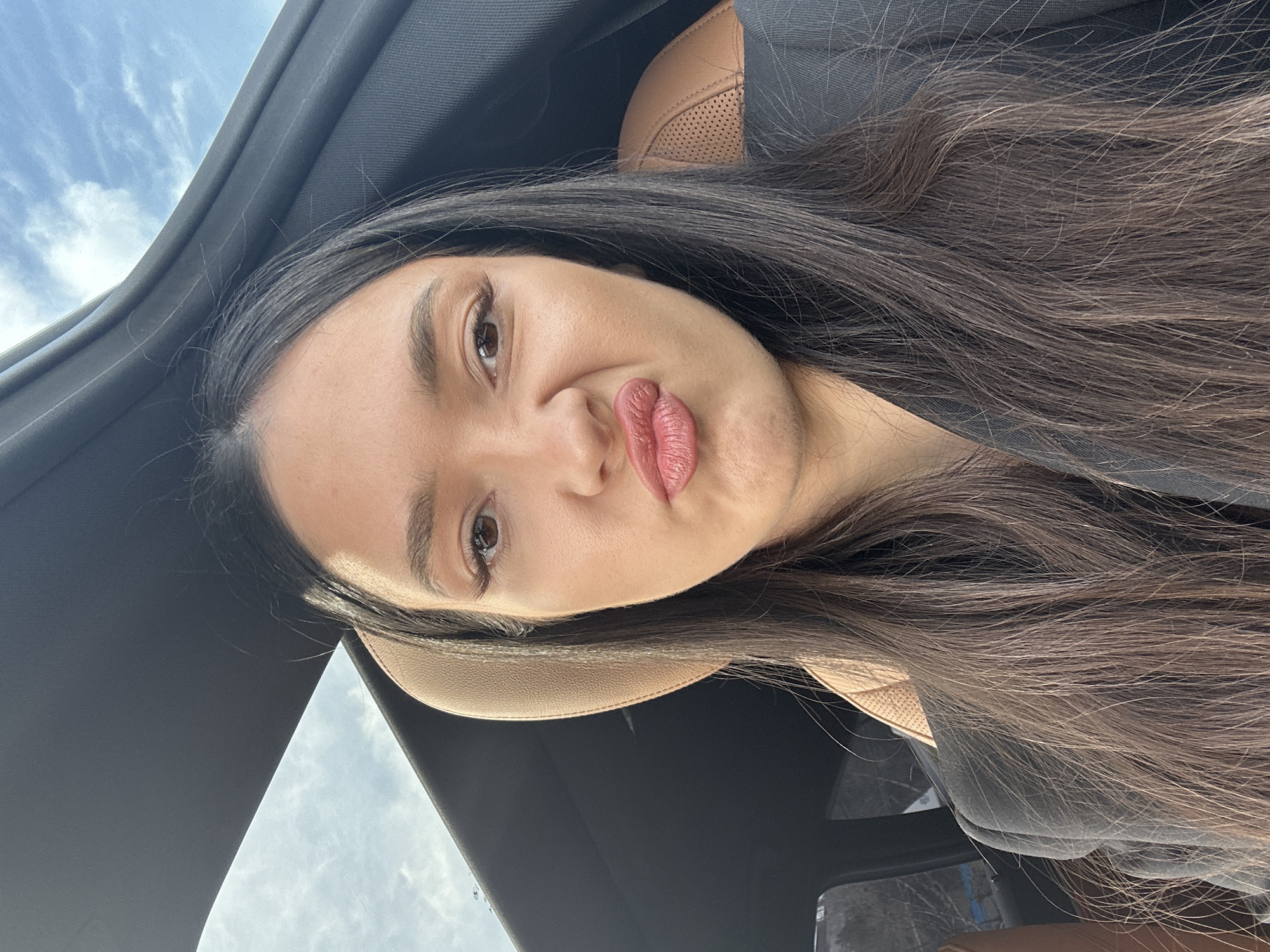 A woman is in a car. The sunroof is open, revealing a cloudy sky. She is wearing a dark jacket and has long, dark hair. She is making a kissing face. The interior of the car has tan leather seats. There is no information to indicate this is in Yerevan, Armenia.